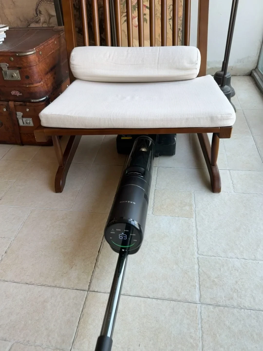 A black Dreame wet-and-dry vacuum cleaner lies flat on a light-colored tiled floor, with its roller head reaching underneath a wooden chair with white cushions.