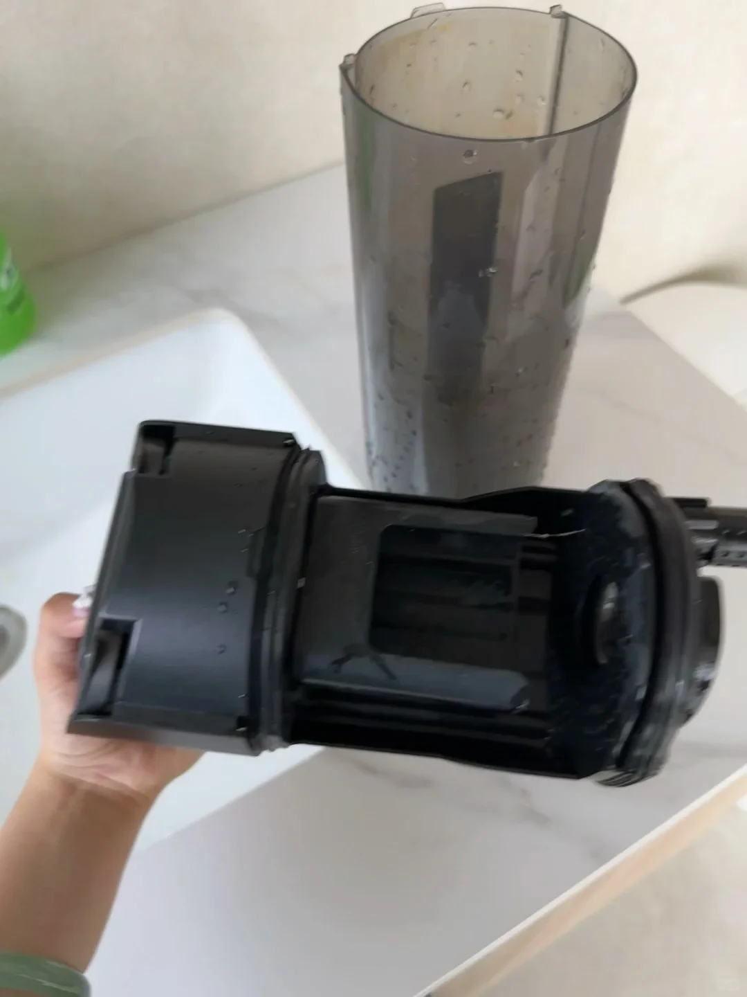 A hand holds a disassembled black plastic appliance assembly above a counter, with a tall, semi-transparent cylindrical tank standing in the background.