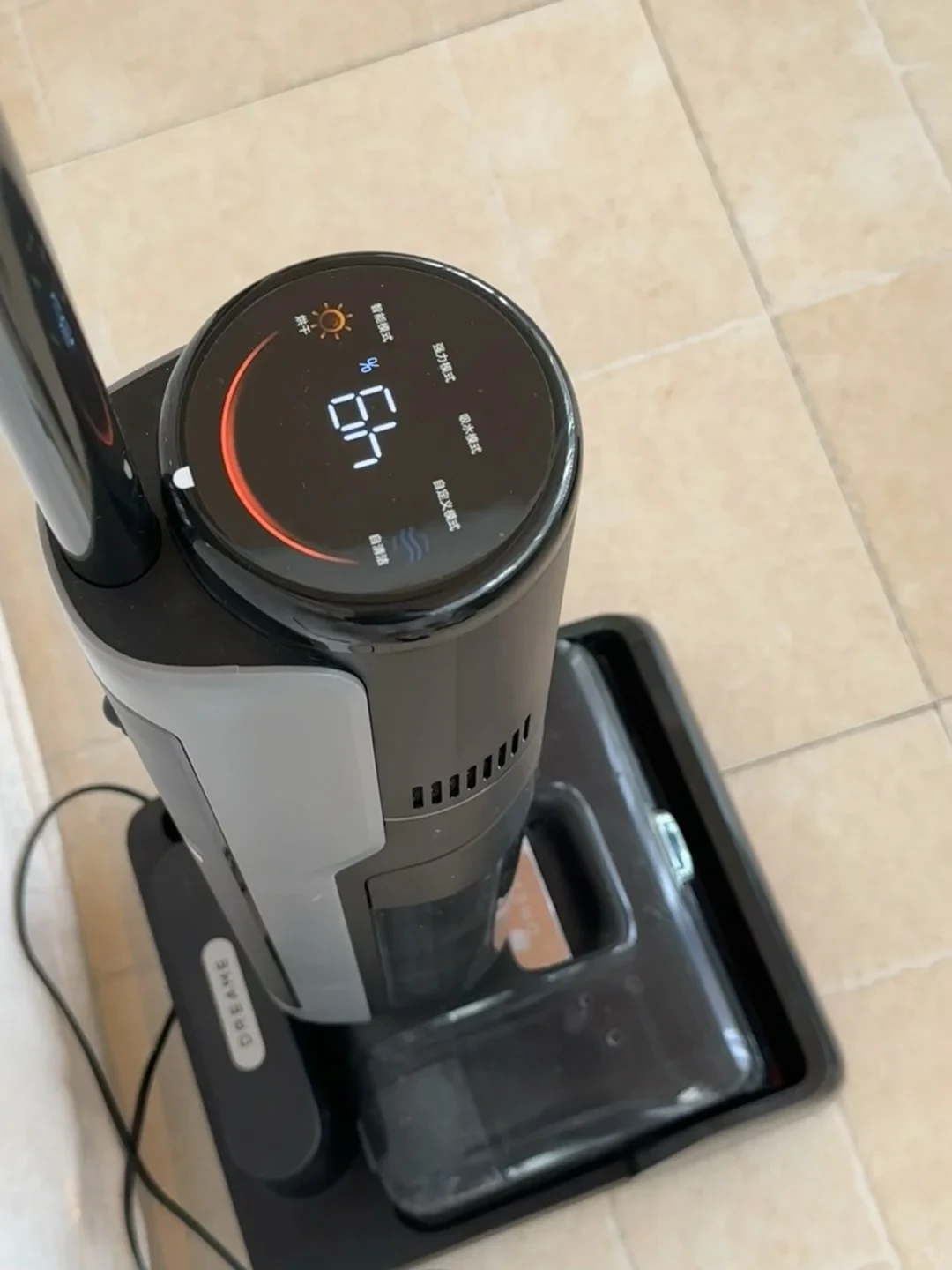 A high-angle view of a Dreame wet-and-dry cordless vacuum cleaner docked on its cleaning base over beige tile flooring. Its round digital top display illuminates 64% battery level, a red charging arc, and control indicators in Chinese.
