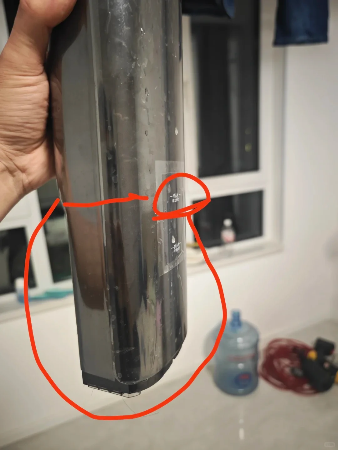 A person holding the translucent dark grey water tank of a floor washer or wet-dry vacuum cleaner. A sticker on the side indicates two maximum fill lines, with a hand-drawn red circle and line highlighting the upper fill line and lower tank body.