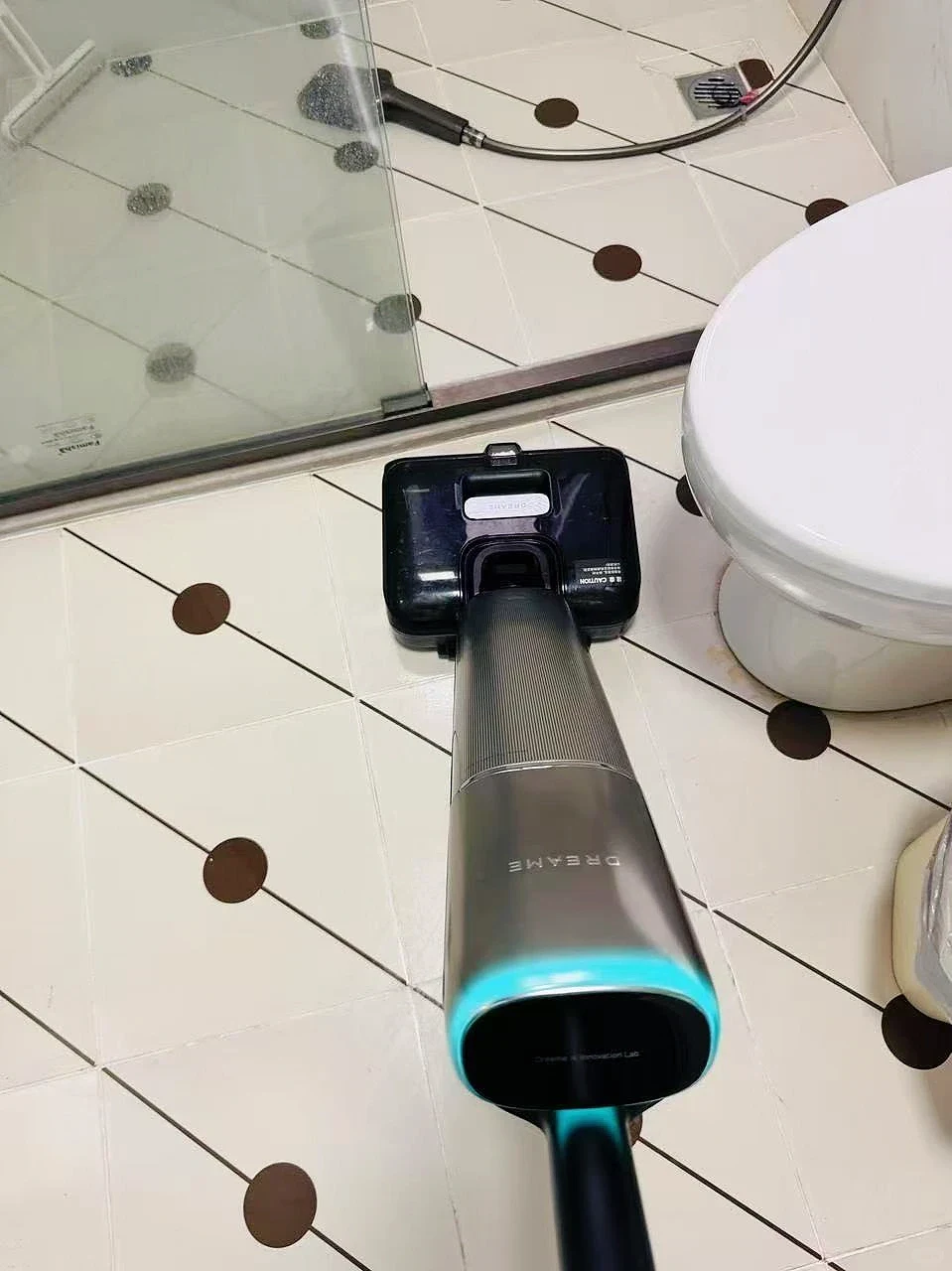 A top-down perspective of a Dreame wet and dry floor vacuum cleaner being used on a white tiled bathroom floor with diamond-shaped lines and brown circular accents, situated between a glass shower enclosure and a white toilet.