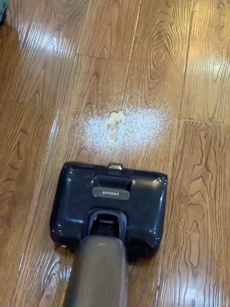 High-angle view of a black Dreame wet-and-dry vacuum cleaner head approaching a spill of white powder and food paste on a wood-grain floor.