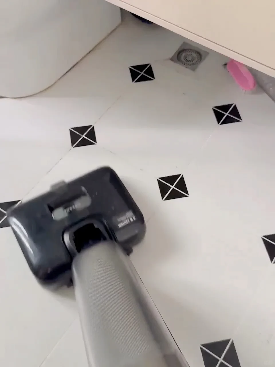 A handheld wet-dry vacuum cleaner head gliding across white tiled bathroom flooring accented with small black geometric corner squares.