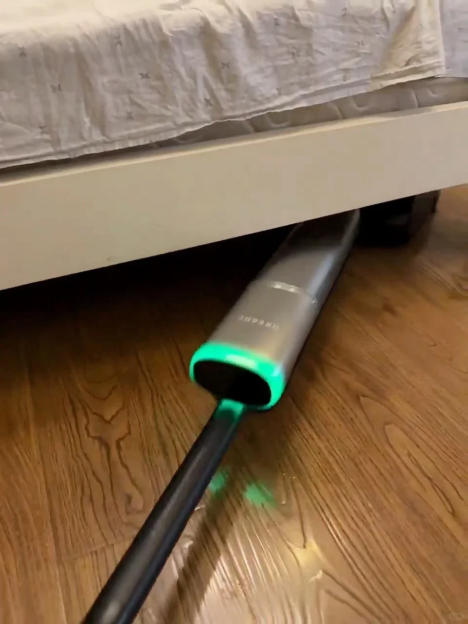 A sleek silver cordless floor cleaner lying flat on a wood floor as it cleans underneath the side of a white bed frame, illuminated with a bright green LED light.