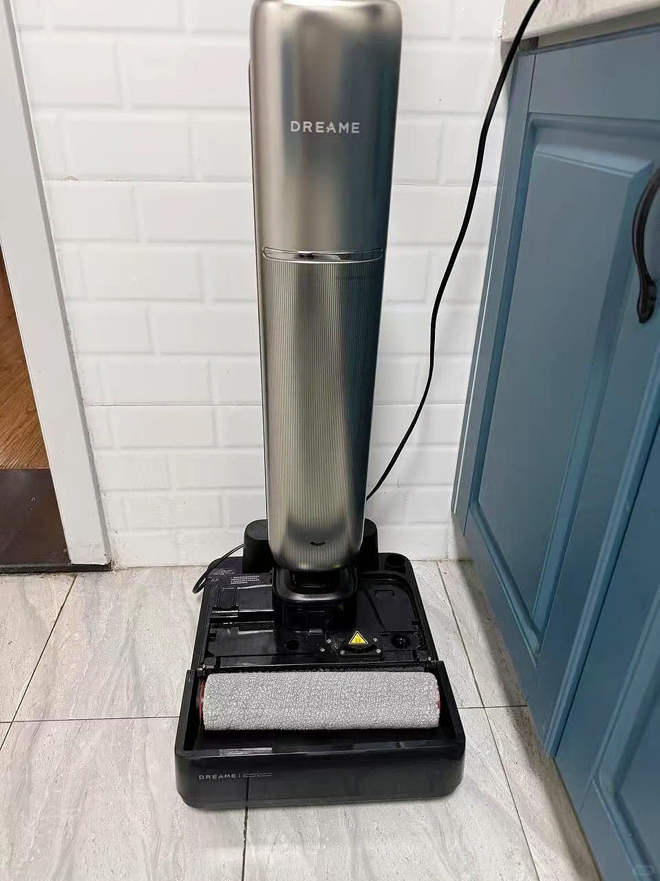 A metallic grey Dreame upright wet and dry floor vacuum cleaner resting on its black base station on a tiled floor next to a blue cabinet.