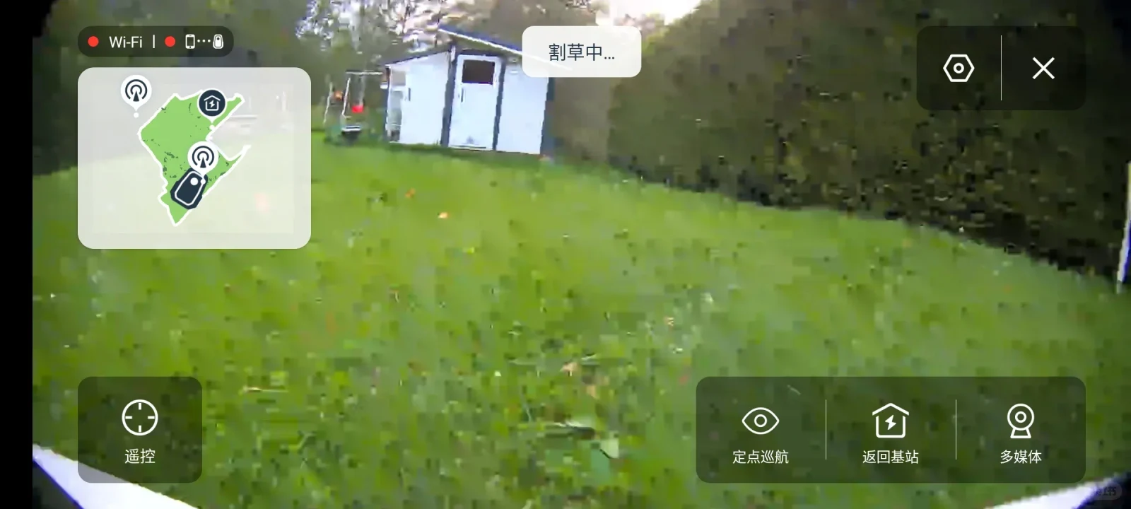 Screen interface displaying a first-person live camera feed from a robotic lawn mower navigating across a grassy backyard. A white garden shed and tall green hedge appear in the background. Interface overlays include an active lawn coverage map on the top left, a status badge reading '割草中...' (Mowing...) at the top center, and navigation buttons at the bottom for remote control, spot cruise, returning to the base station, and multimedia.