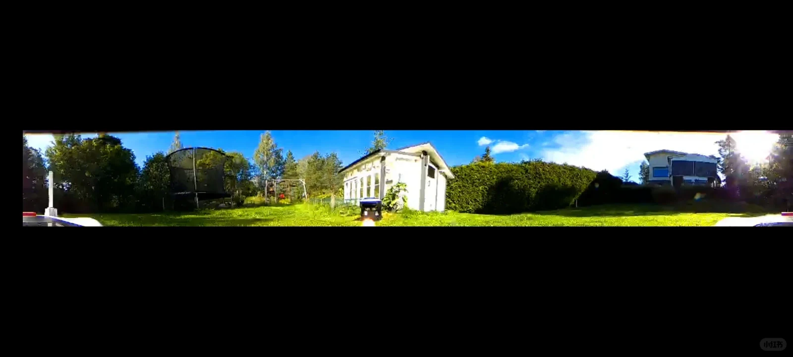An ultra-wide panoramic view of a sunny backyard with green grass, a trampoline with a safety enclosure on the left, a white shed or small outbuilding in the center, and trees, hedges, and a house on the right under a bright blue sky.
