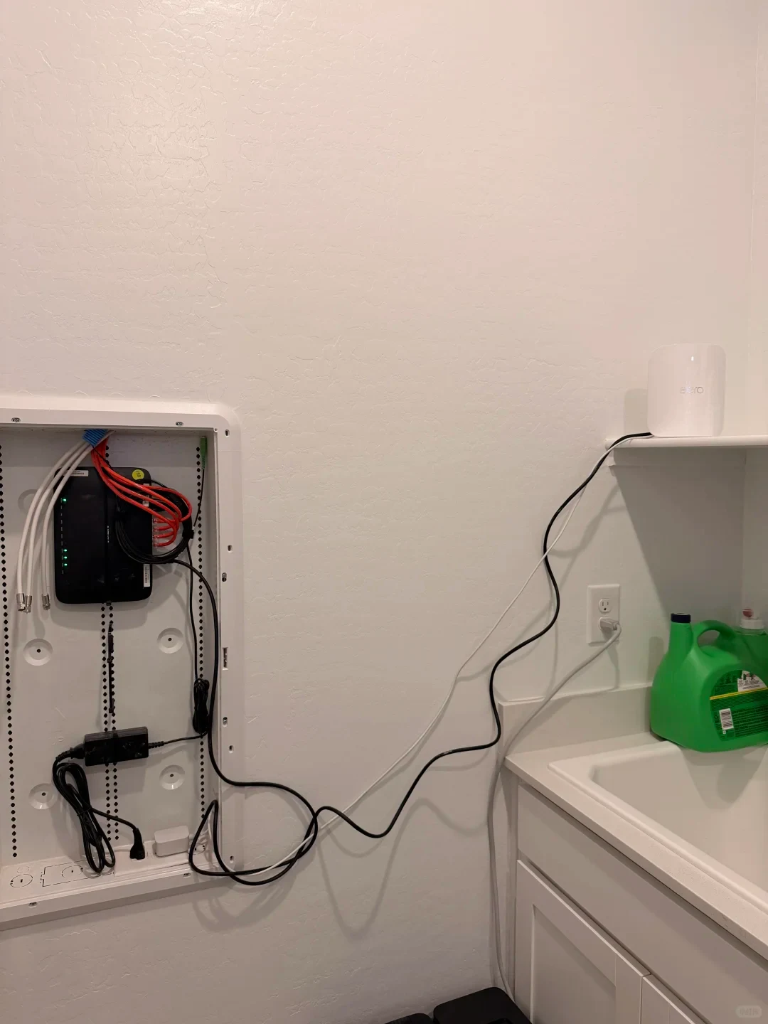 An open in-wall media enclosure containing networking equipment, with cables extending out across a wall to a white eero router placed on a shelf above a utility sink.