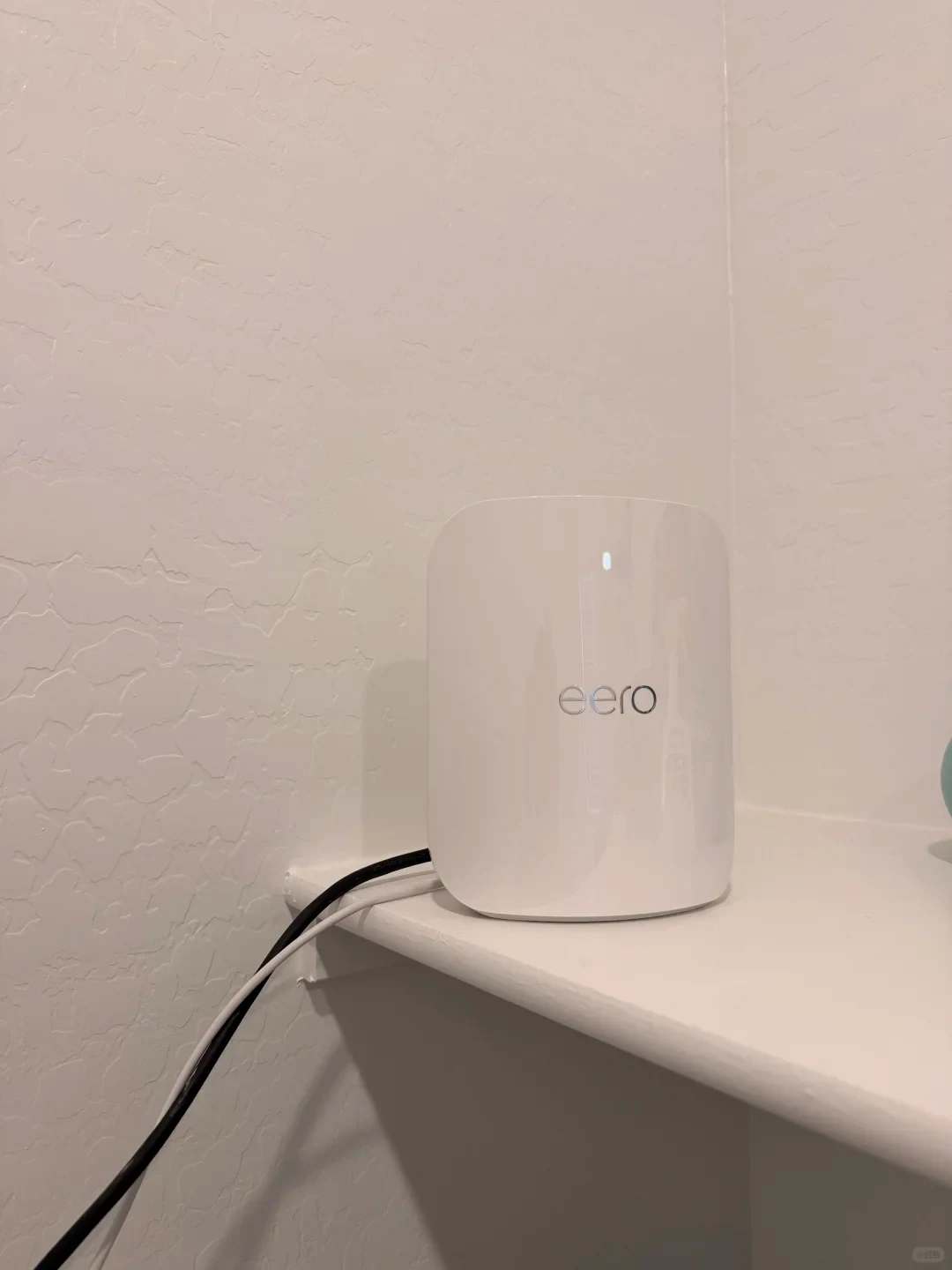 A white eero Wi-Fi router sitting on a white shelf near a textured wall, connected with black and white cables.