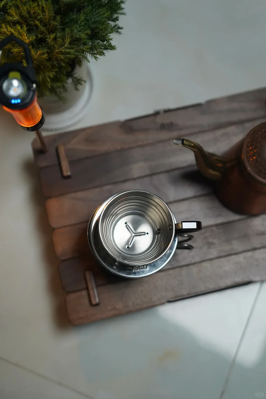 A stainless-steel Kalita dripper sits over a dark mug, showing three drain holes and a raised three-pronged pattern in its flat base. A gooseneck kettle and lantern stand nearby.