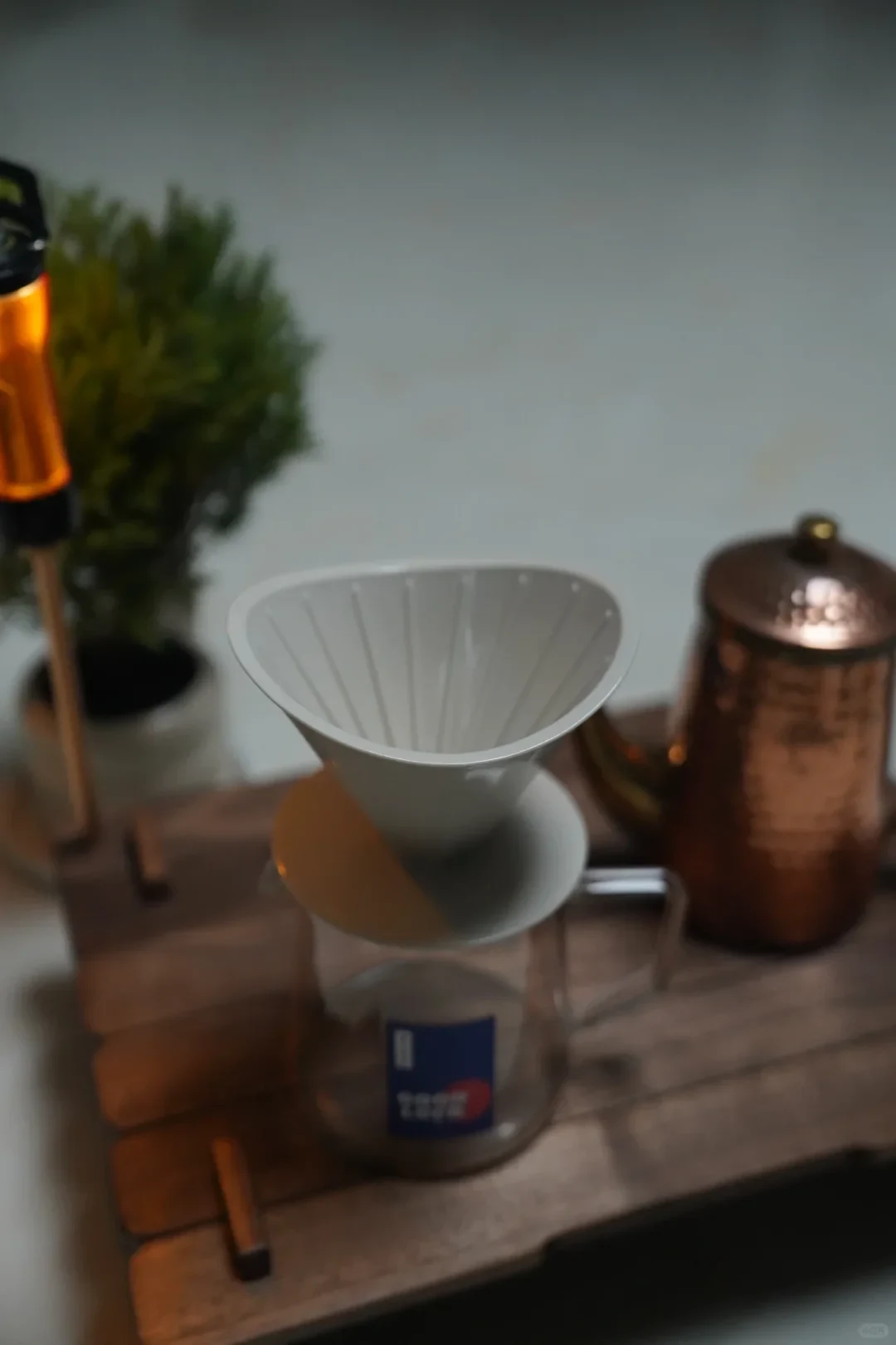A white ribbed pour-over coffee dripper rests on a clear glass server on a wooden tray, with a copper-colored lidded coffee pot and a small potted plant nearby.