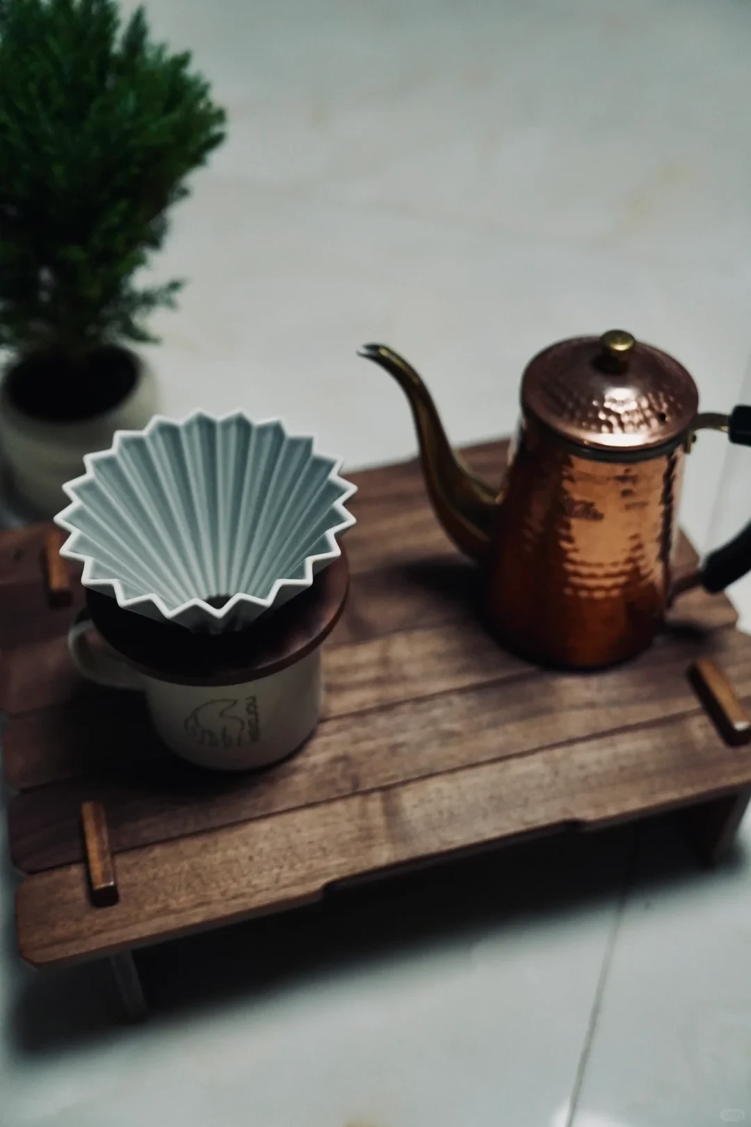 A grey ceramic ORIGAMI dripper sits in a wooden holder over a mug beside a copper-colored gooseneck kettle.