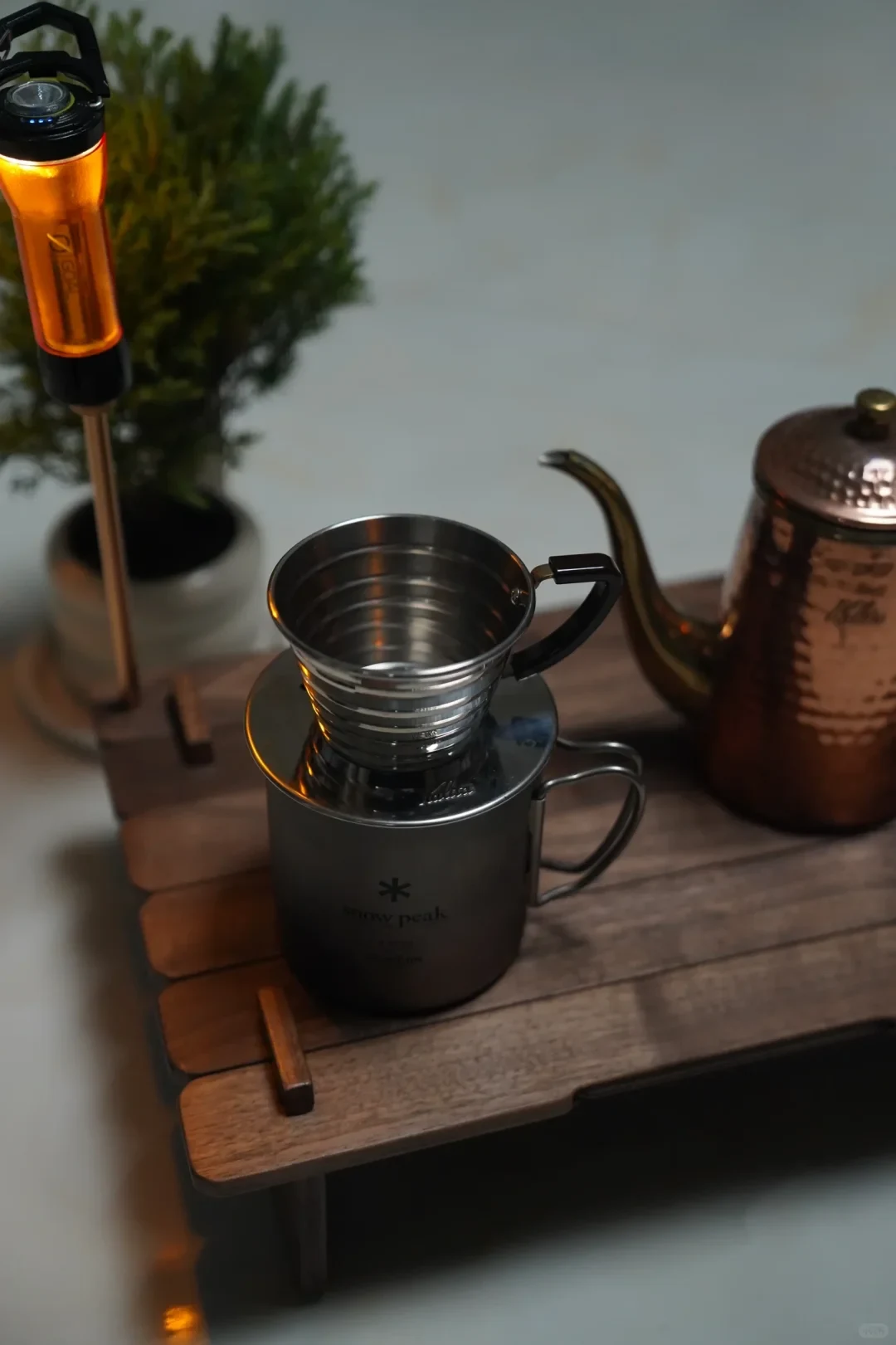 A stainless-steel coffee dripper sits on a dark camping mug on a wooden table, with an orange lantern to the left and a copper-colored gooseneck kettle to the right.