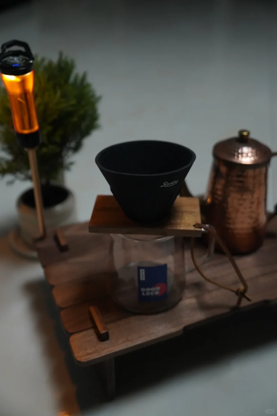 A black cone-shaped coffee dripper sits on a wooden stand above a clear glass server. A glowing orange lantern is on the left, and a copper-colored kettle is on the right.