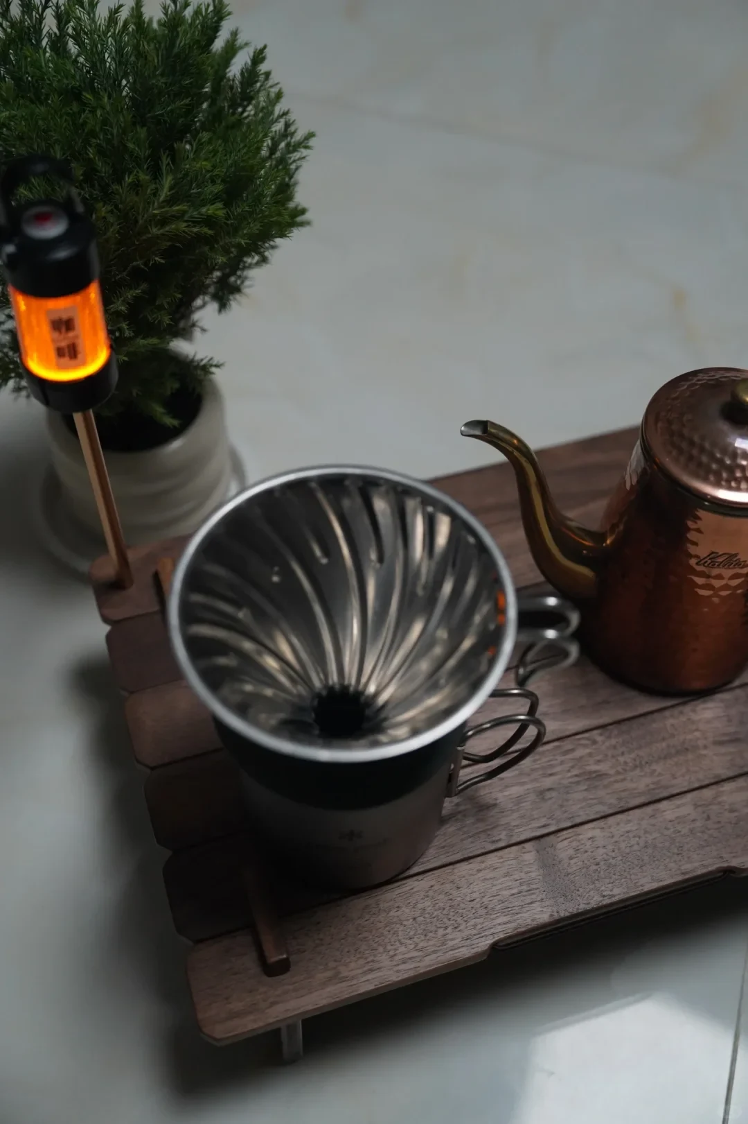 A metal pour-over coffee dripper sits on a wooden slatted stand beside a copper-colored gooseneck kettle. A small orange-lit device stands to the left, with a potted evergreen plant behind it.