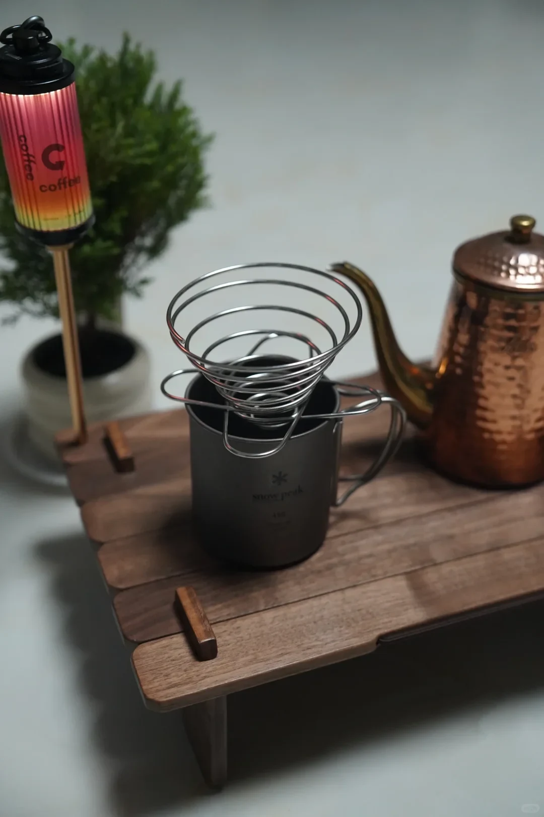 A metal spiral pour-over coffee dripper rests on a dark mug on a wooden camping table, with a copper kettle and a small coffee-themed lamp beside it.