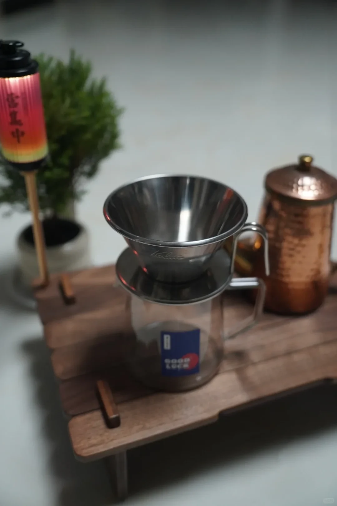 A stainless-steel pour-over coffee dripper sits on a clear glass server on a wooden stand, with a copper-colored lidded vessel and a small lantern nearby.