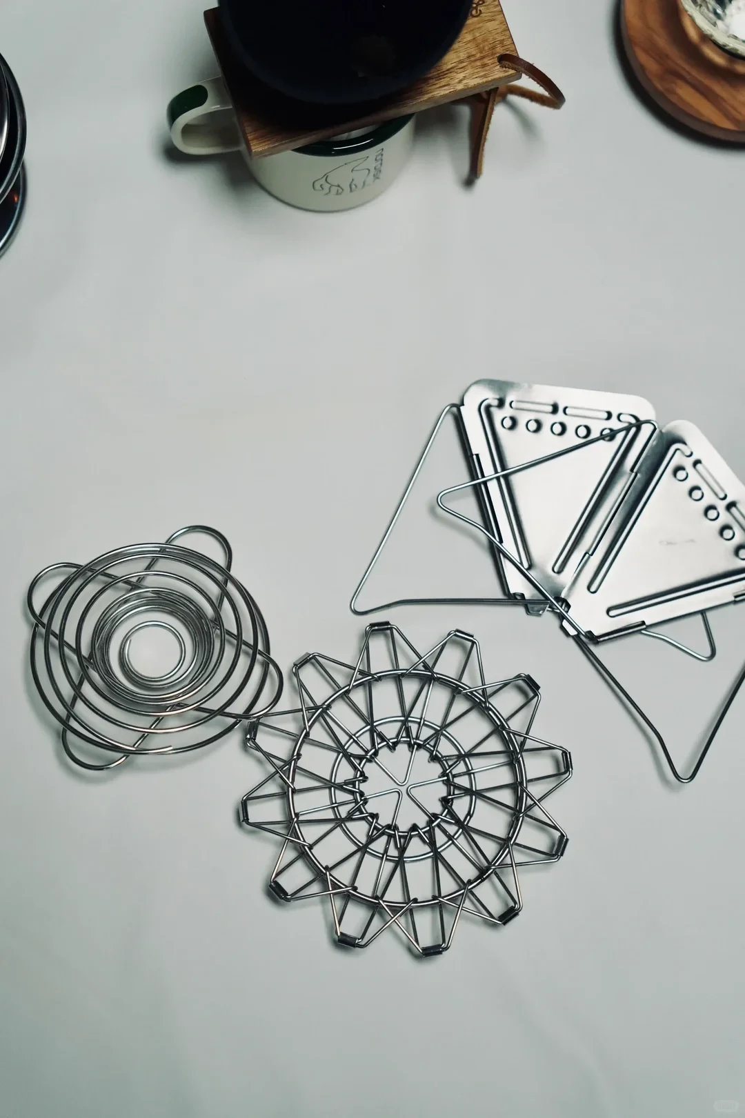 Three folding coffee drippers lie flattened on a pale tabletop: a spiral coil, a hinged wire design and triangular metal plates. A mug and wooden holder are partly visible above them.