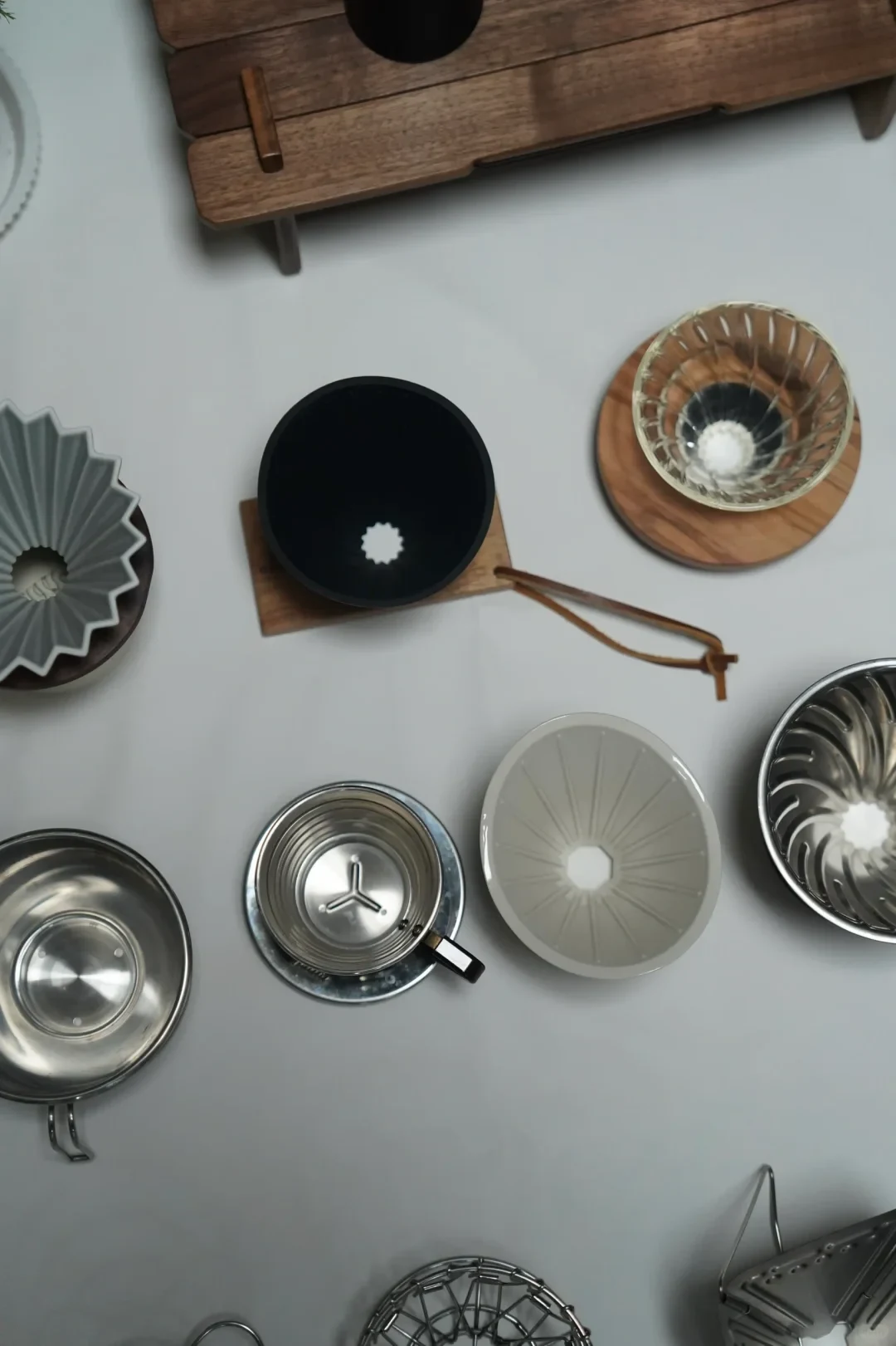 An overhead arrangement of coffee-brewing equipment on a pale surface, including metal and ceramic drippers, a black circular brewer on a wooden base, a glass cup on a wooden coaster, and part of a wooden rack.