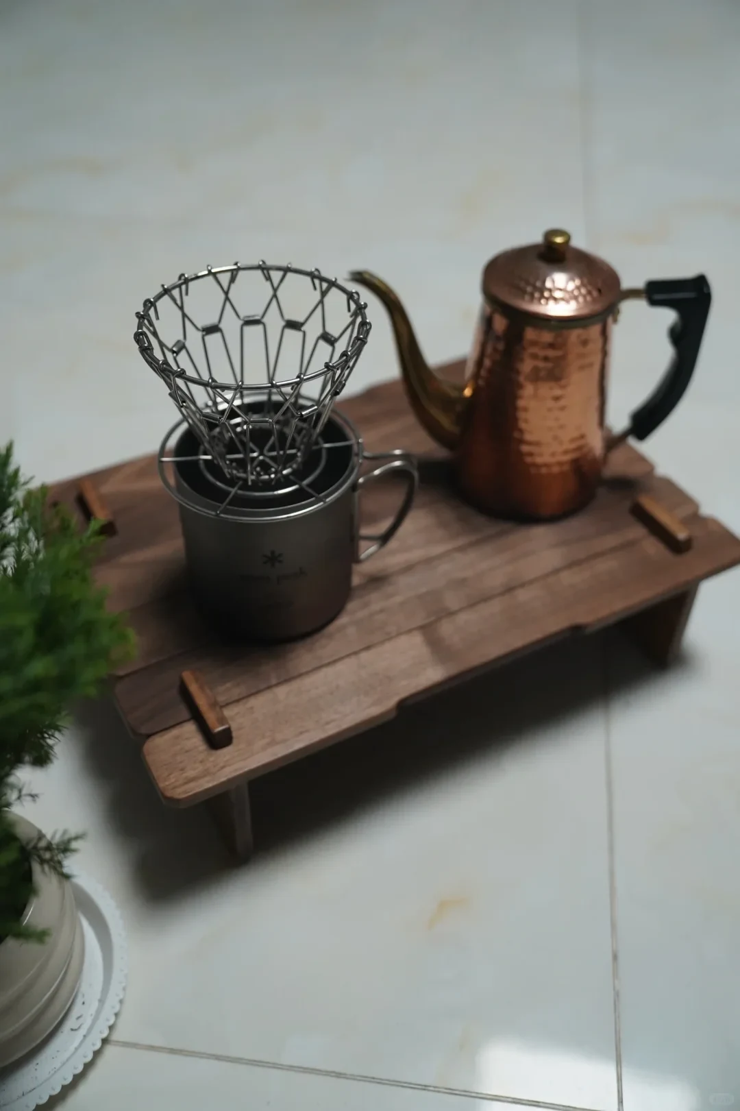 A metal pour-over coffee dripper sits on a dark mug on a wooden slatted tray, beside a copper-colored gooseneck kettle. Part of a potted plant is visible at the left.