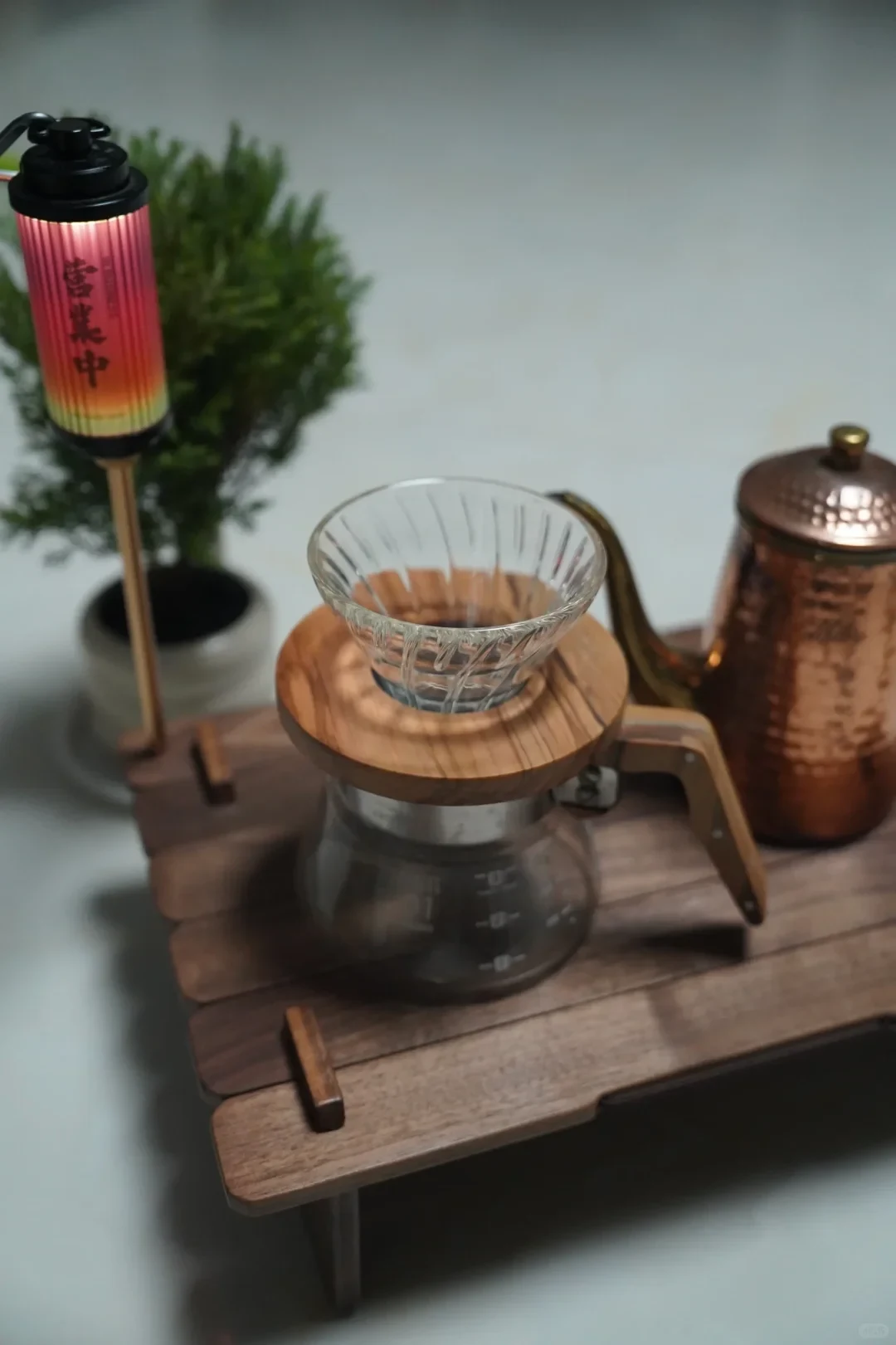 A glass pour-over coffee dripper sits on a wooden ring above a marked glass carafe on a small wooden stand. A copper kettle and a red lantern-shaped light are beside it, with a potted plant in the background.
