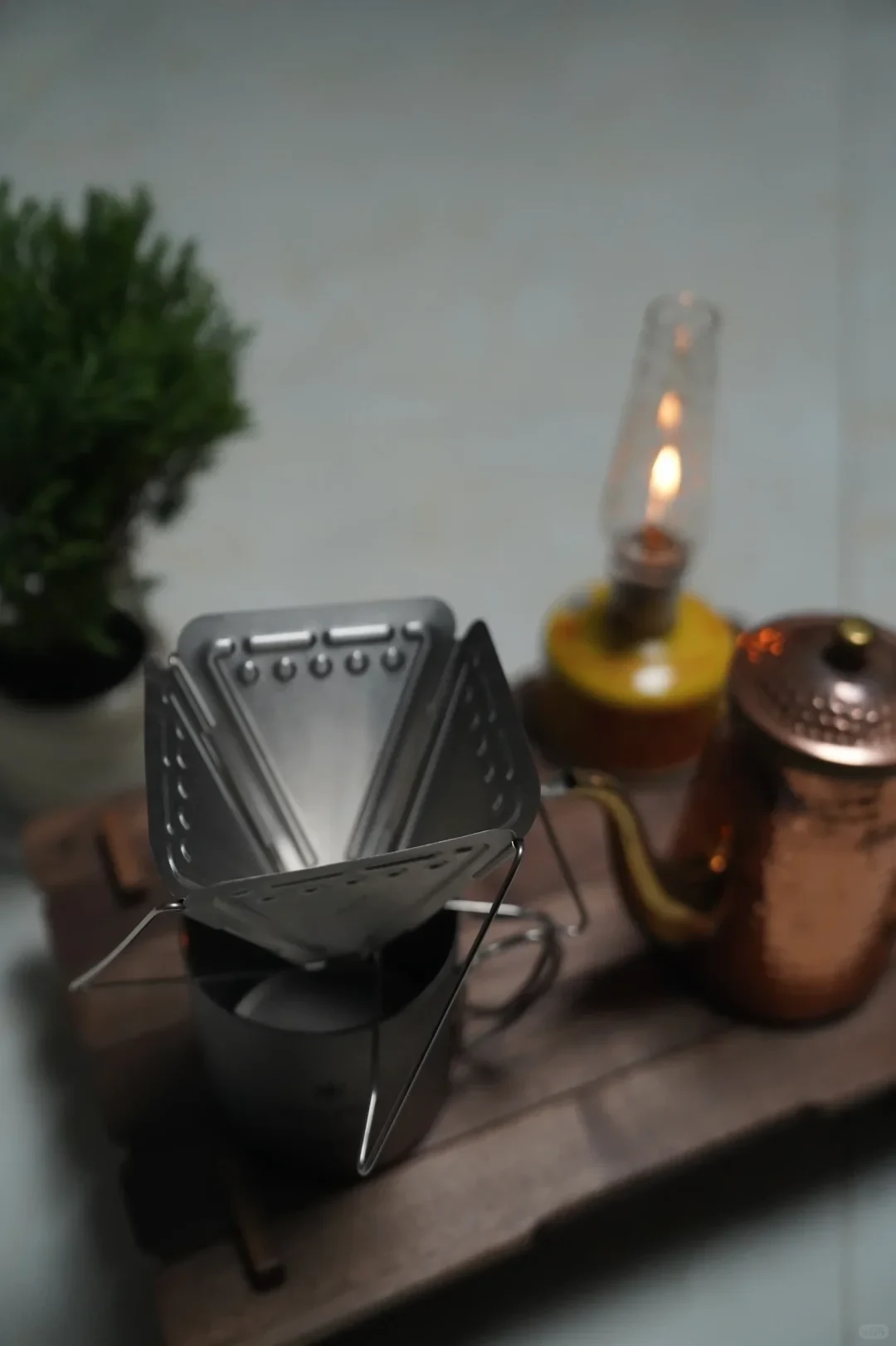 An open triangular folding coffee dripper stands on a dark mug beside a glass lamp and a copper-colored gooseneck kettle. The perforated metal plates form the dripper walls.