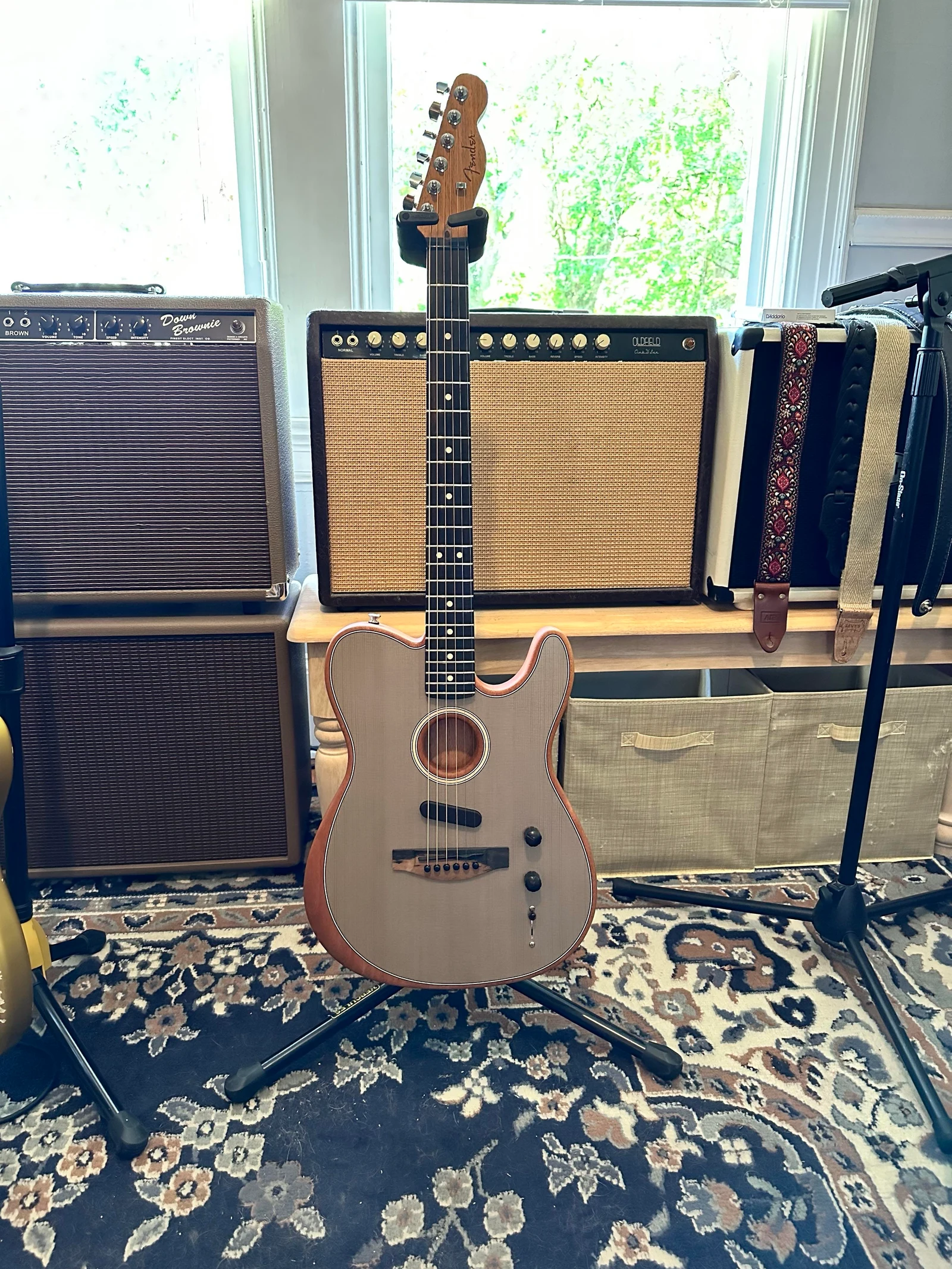 A natural-finish Telecaster-shaped Acoustasonic on a stand among amplifiers.