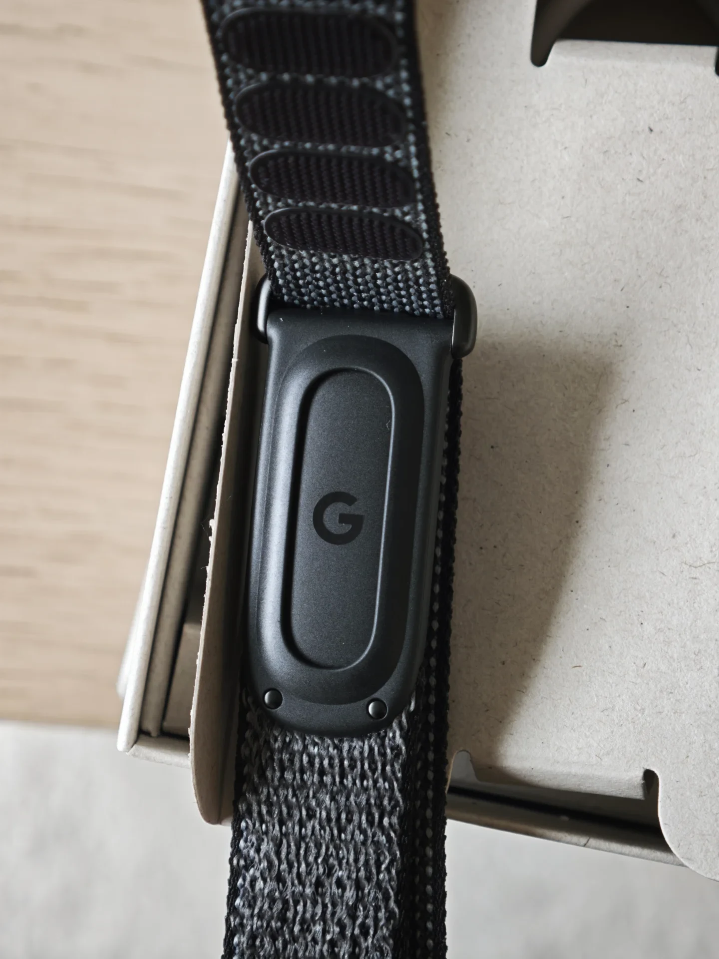 A close-up shot of a black oblong Google accessory featuring the letter G logo, attached to a dark grey woven loop strap inside cardboard packaging.