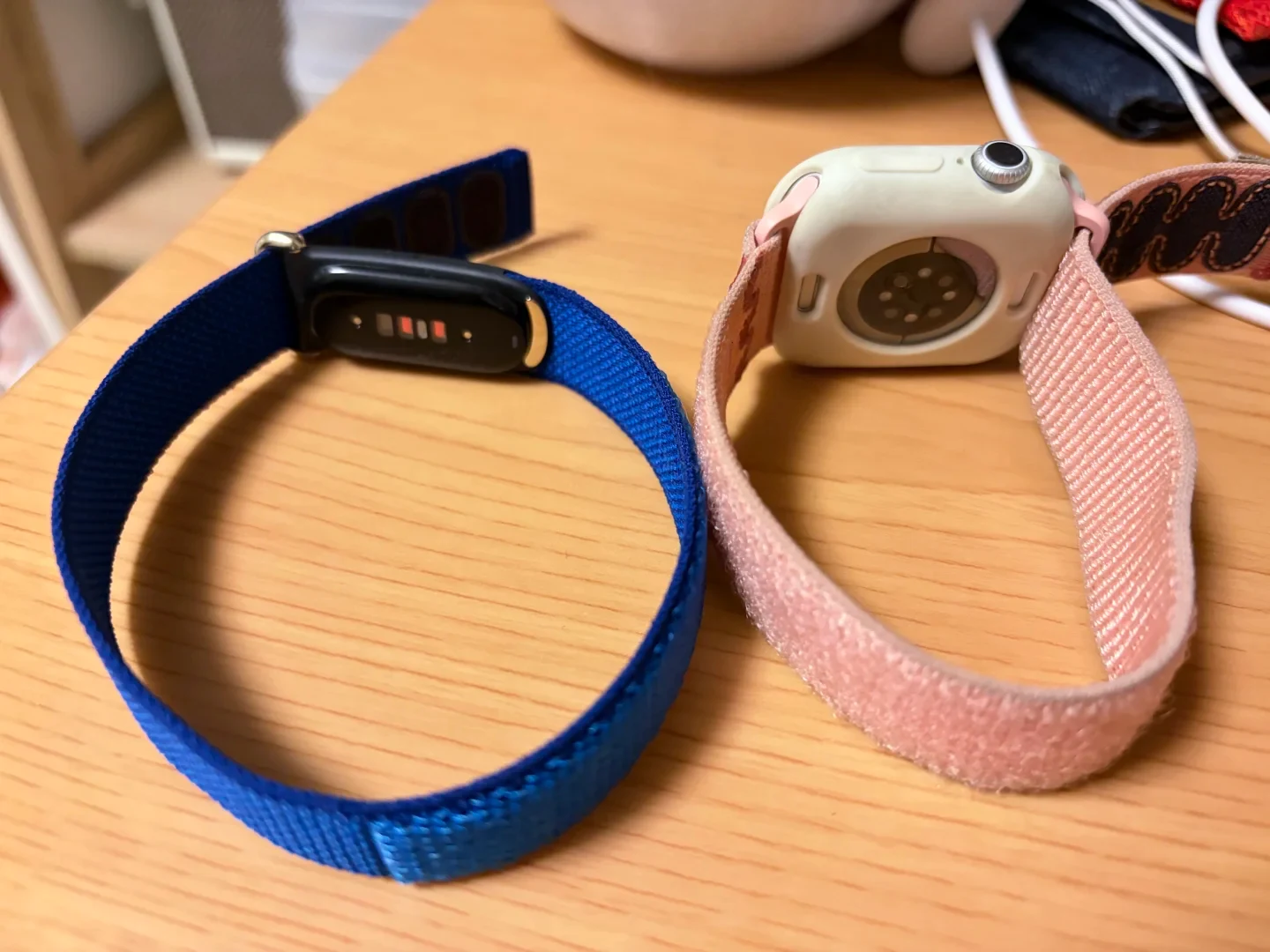 A fitness tracker with a blue nylon loop band alongside a smartwatch with a protective case and pink fabric loop band, resting on a wooden surface.