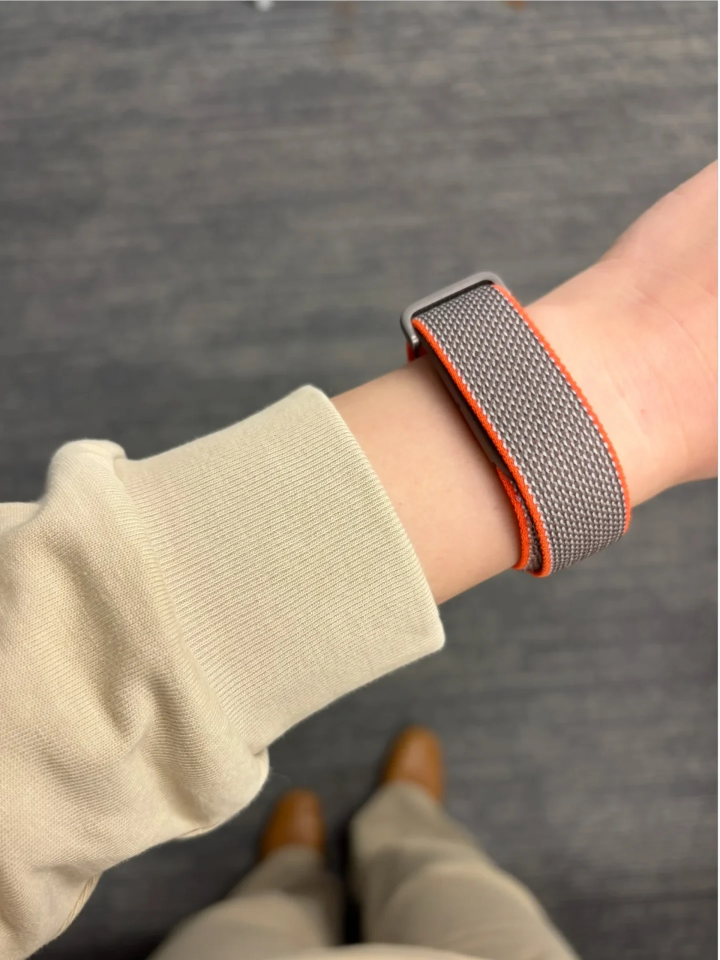 A close-up view of a person's wrist wearing a gray woven nylon smartwatch strap with bright orange edges, paired with a beige long-sleeve cuff against a dark carpeted floor.