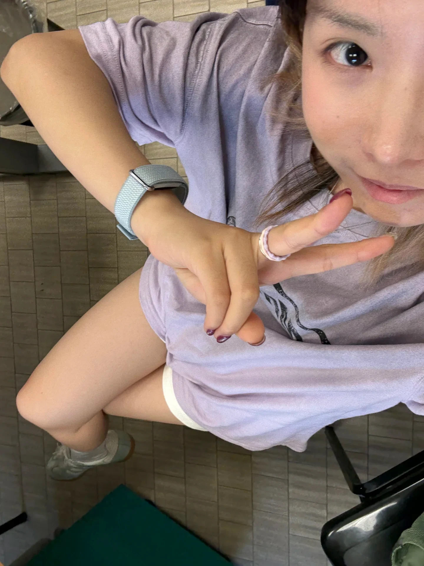 High-angle selfie of a woman wearing an oversized lavender T-shirt and white shorts, sitting down and flashing a peace sign toward the camera.