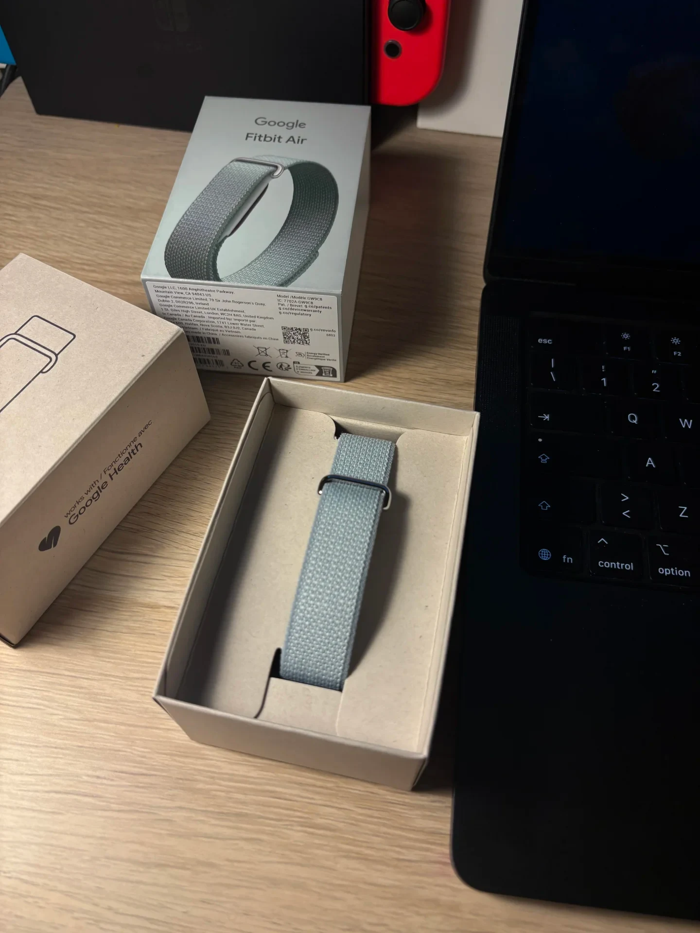 An unboxed Google Fitbit Air woven strap resting in its cardboard tray on a desk beside its packaging boxes and a laptop keyboard.