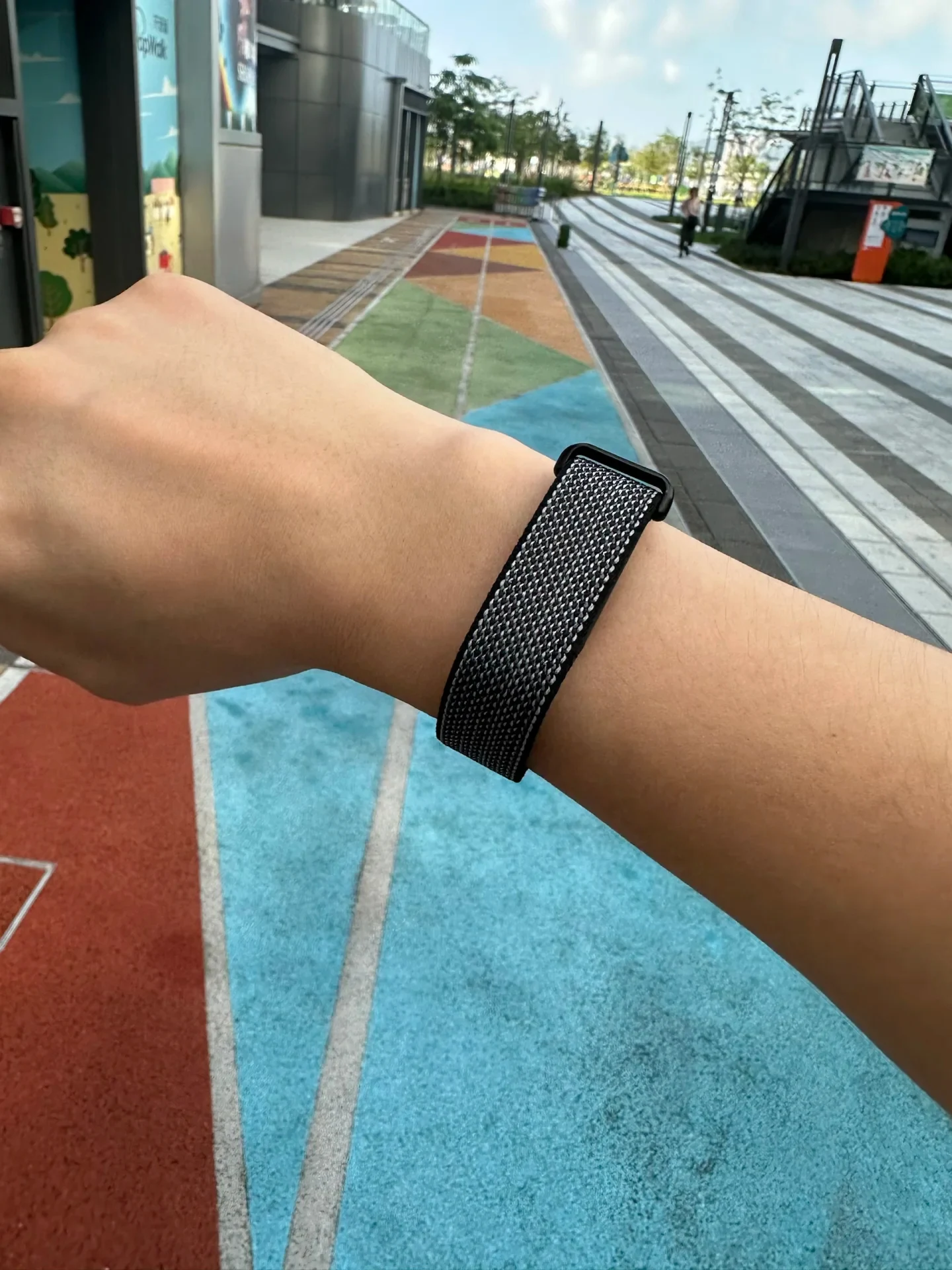 Close-up of a person's wrist wearing a smartwatch secured with a black and gray woven nylon strap, photographed outdoors above a colorful running track.