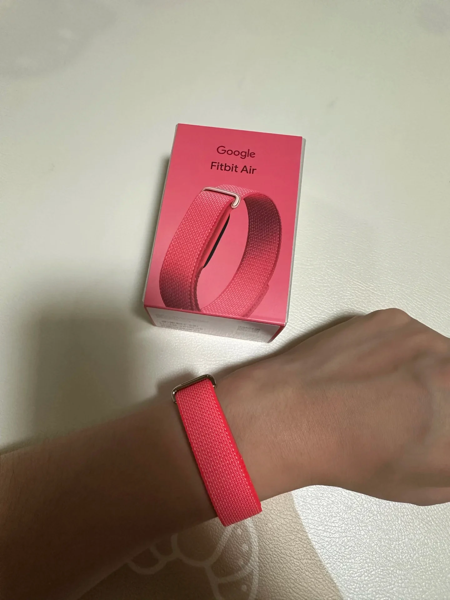 A person's wrist wearing a pink woven strap, shown next to a pink product box labeled "Google Fitbit Air" featuring an illustration of the strap.