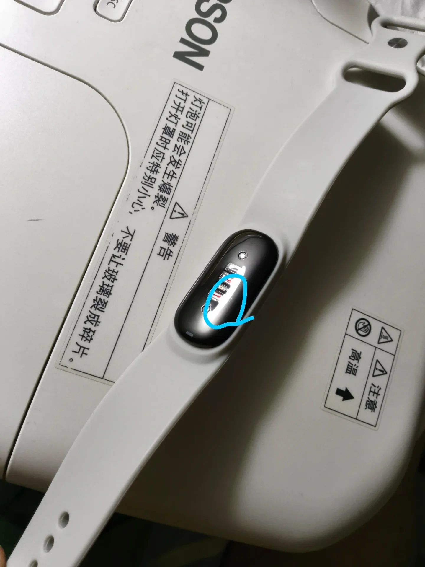A smart fitness tracker with a white silicone strap resting on a white device casing, featuring a hand-drawn blue circle highlighting a small blemish on its curved glass screen.