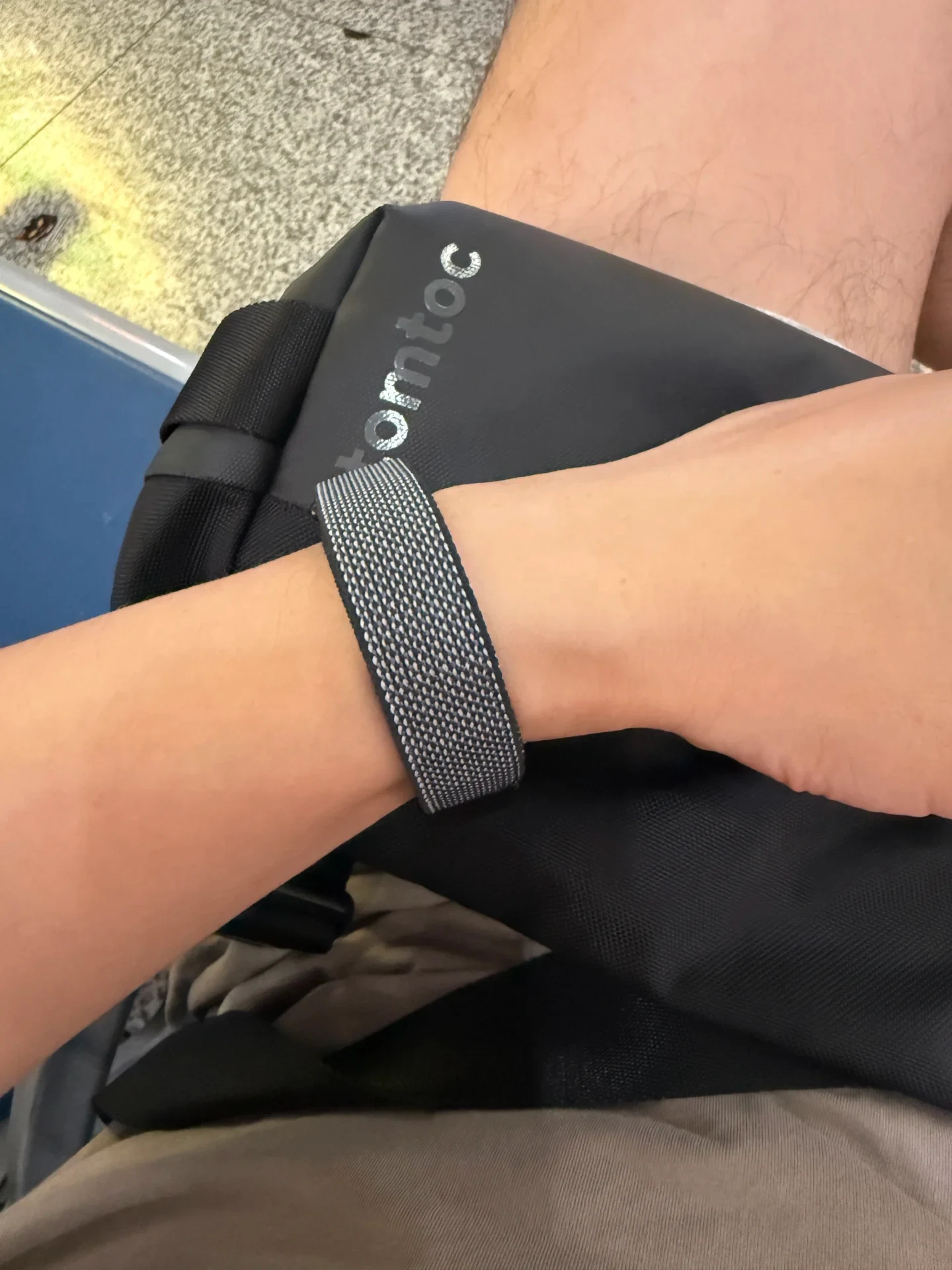 A close-up of a person's wrist wearing a gray woven fabric strap, resting over a black tomtoc bag placed on their lap.