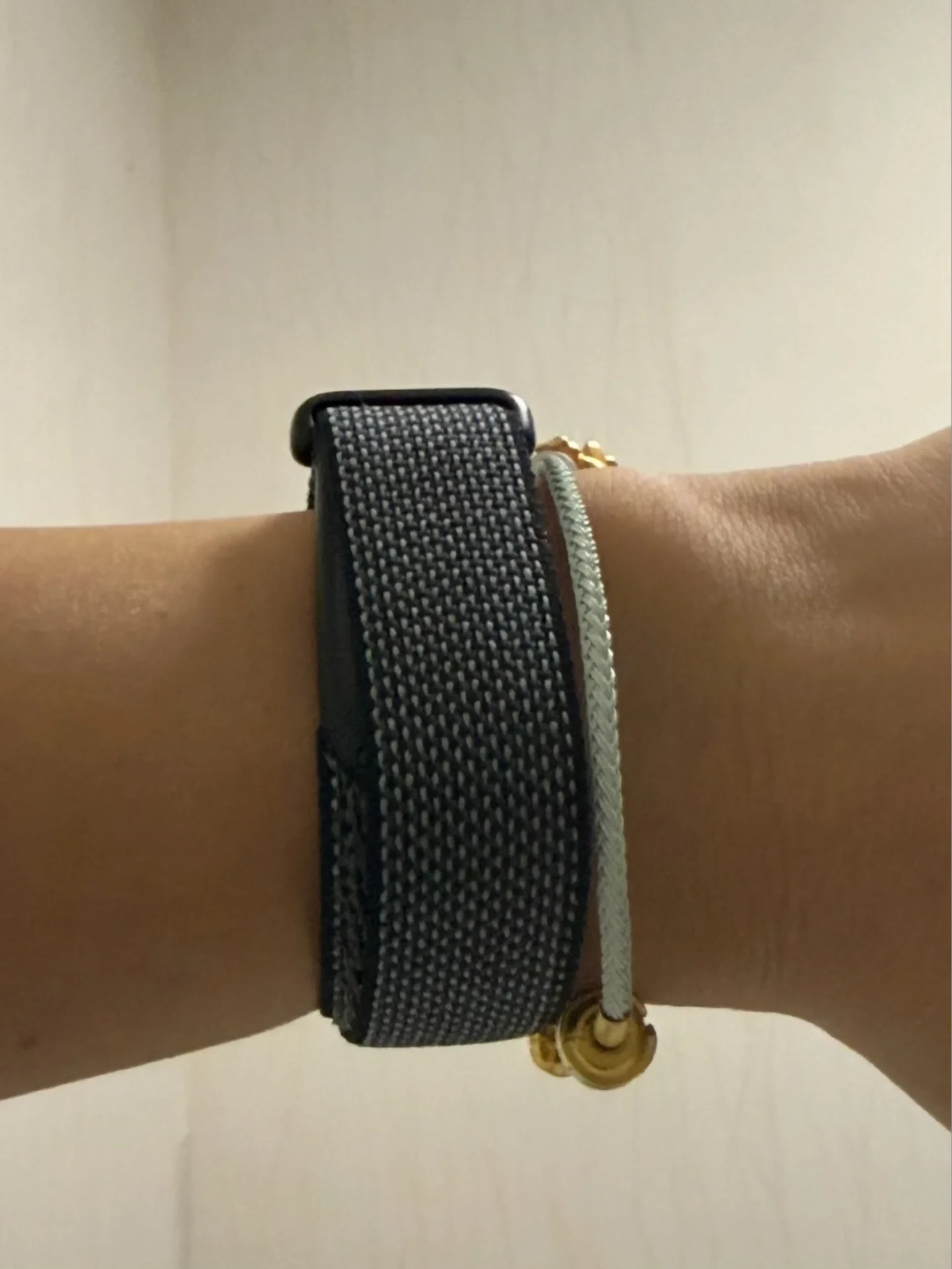 Close-up of a person's wrist wearing a dark gray woven nylon loop smartwatch strap alongside a thin light-colored cord bracelet with gold beads.