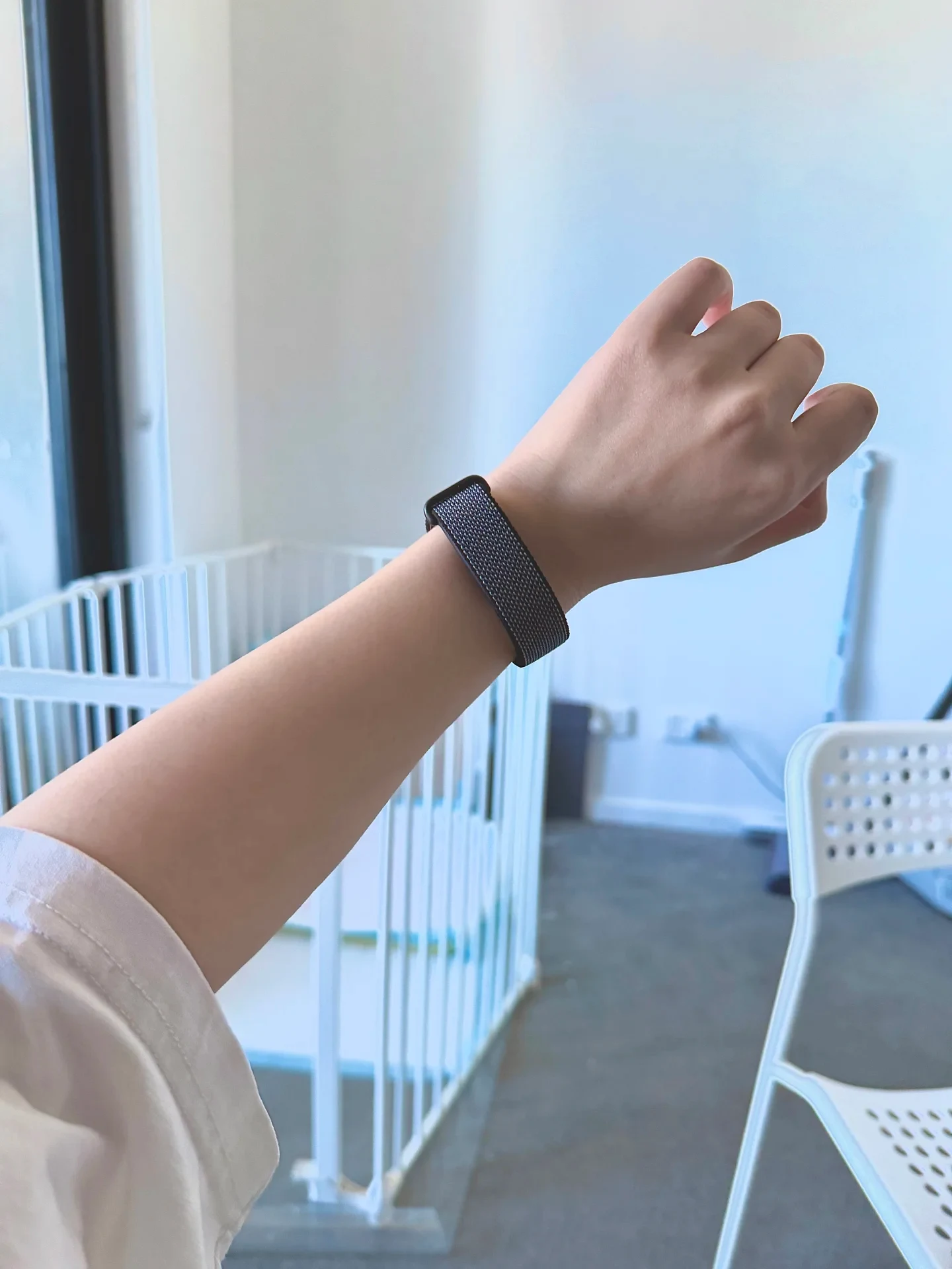 An arm raised showing a dark woven strap of a smartwatch or fitness tracker on the wrist, set against an indoor background featuring a white playpen and a chair.