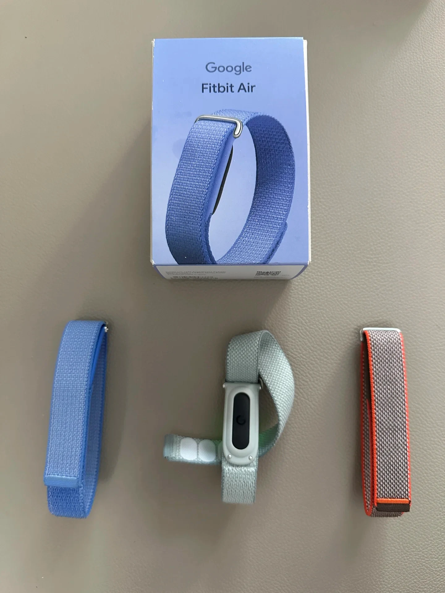 Top-down view of a Google Fitbit Air box along with three woven replacement straps: a blue strap on the left, a light sage strap with a fitness tracker capsule attached in the middle, and a gray strap with orange trim on the right.