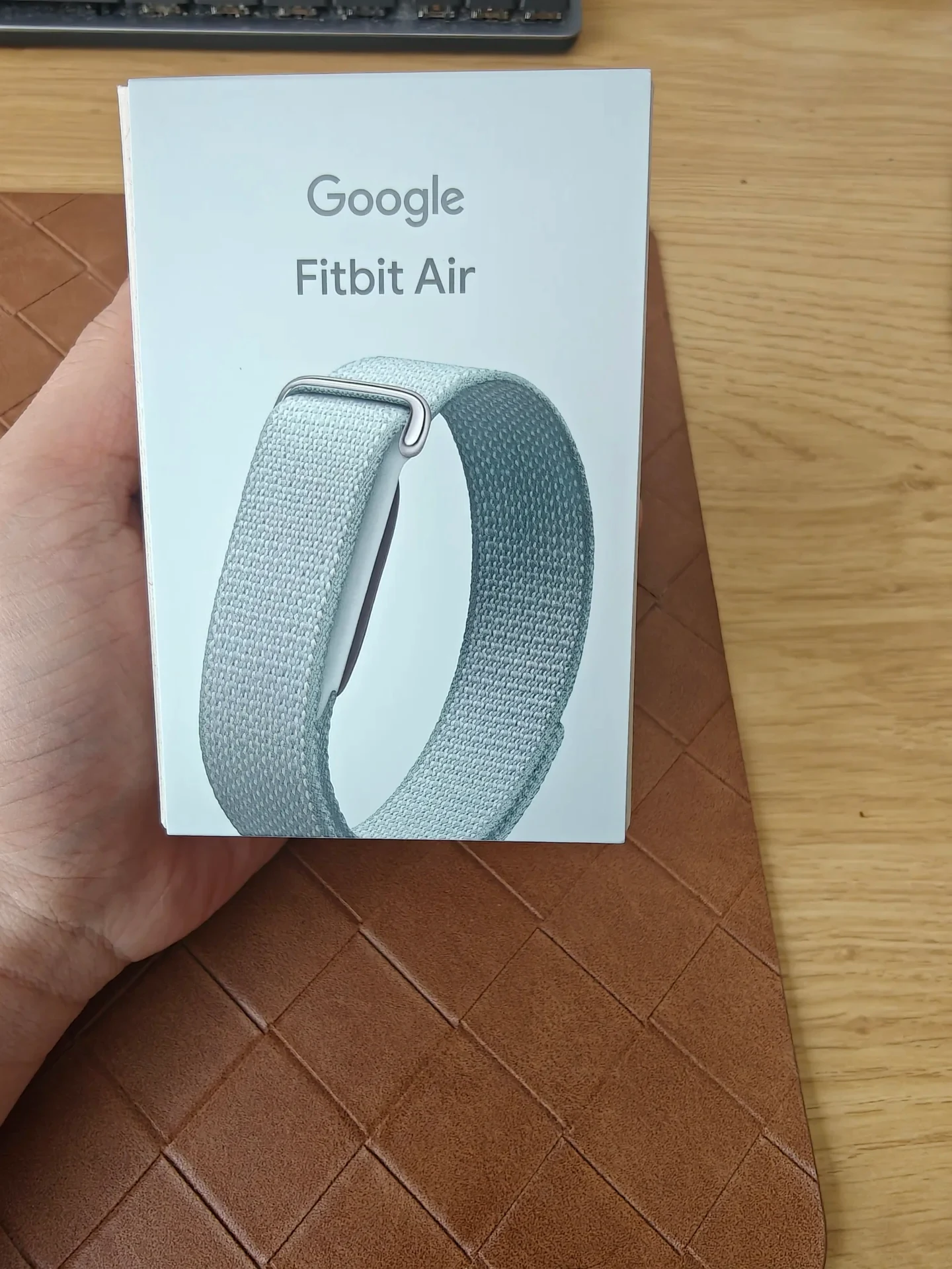 A hand holding the white retail box for a Google Fitbit Air device featuring an image of the fitness tracker with a light blue woven loop band.