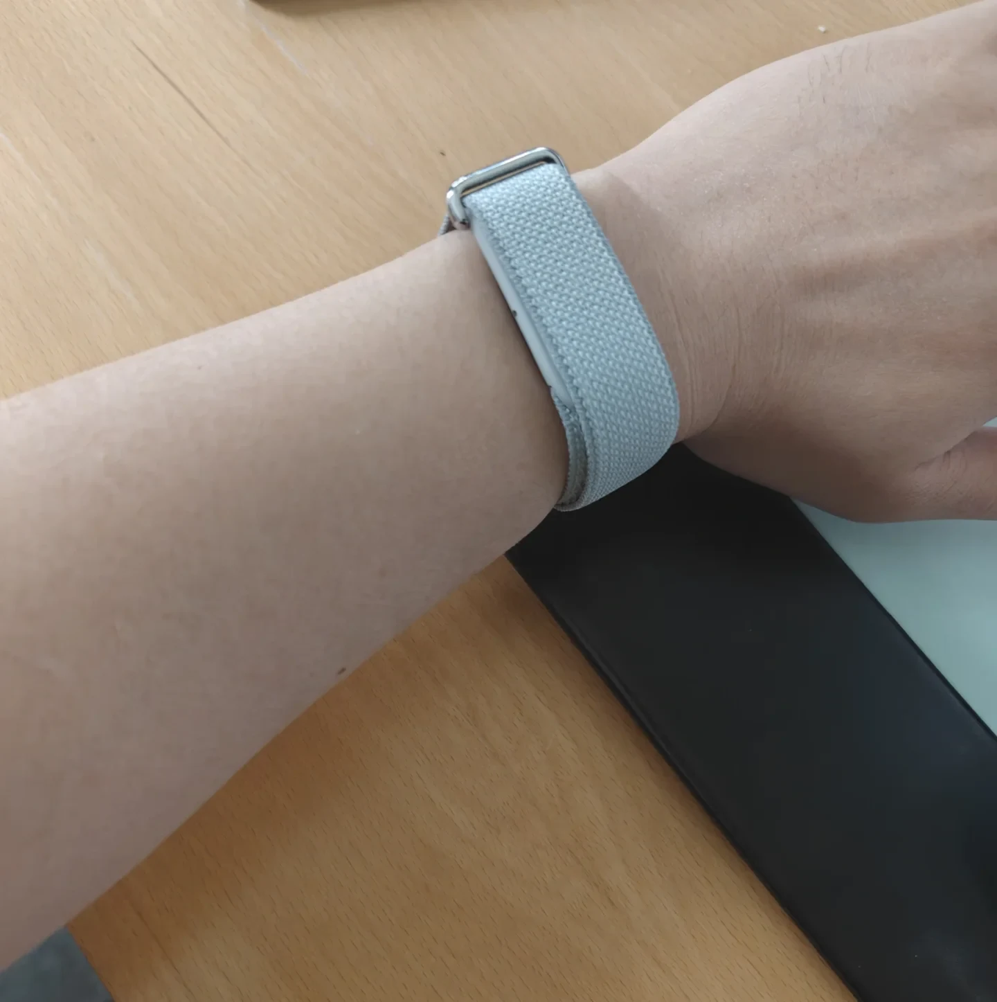Close-up of a person's wrist wearing a light blue woven nylon smartwatch strap with a metal buckle, resting on a wooden desk.