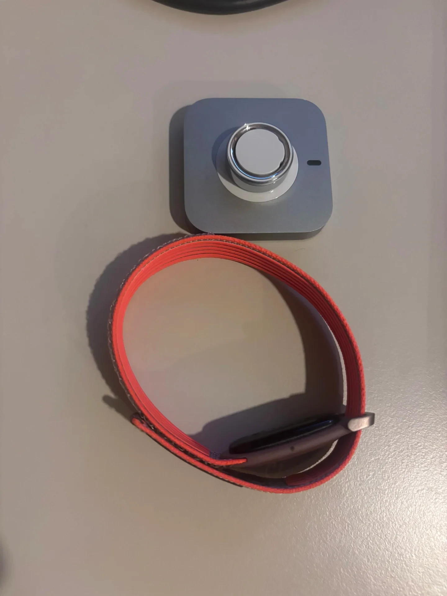Top-down view of a metallic square smartwatch charging base next to a smartwatch with a bright coral-red woven strap on a plain surface.