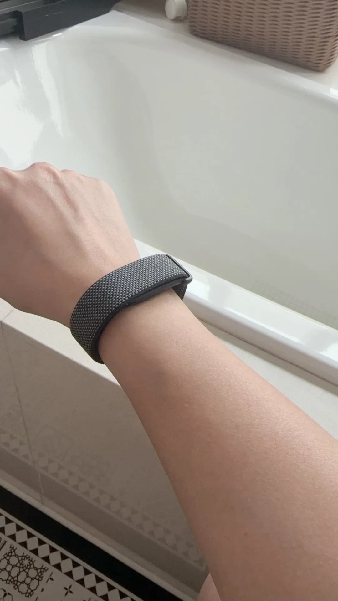 A person's wrist wearing a dark grey woven nylon loop band, positioned against the rim of a white bathtub.