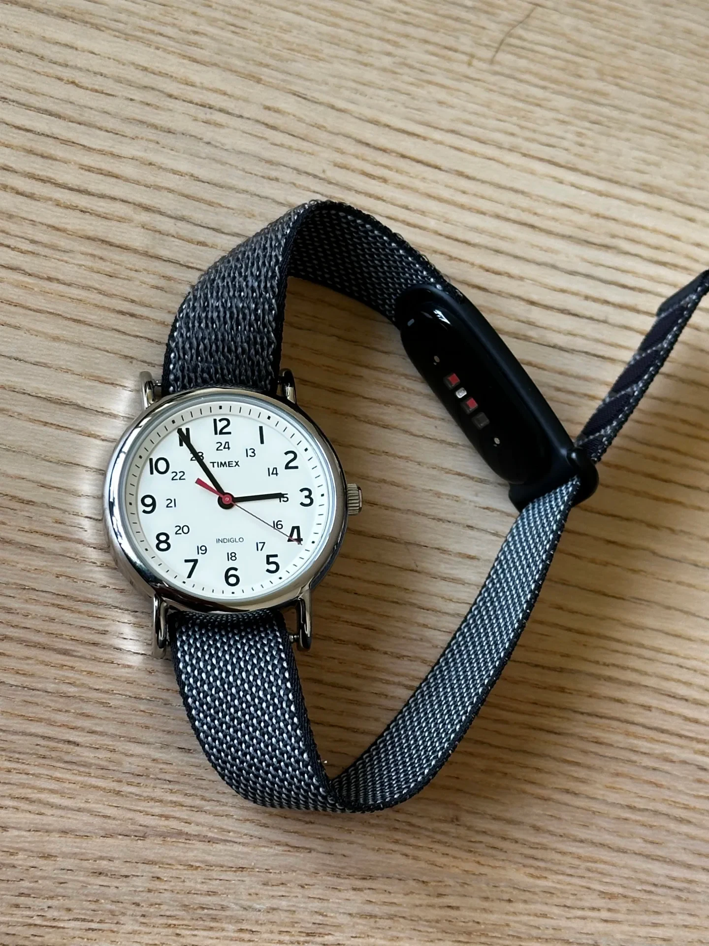 A Timex analog wristwatch paired on a single grey woven strap with a black fitness tracker pill, displayed on a light wood surface.