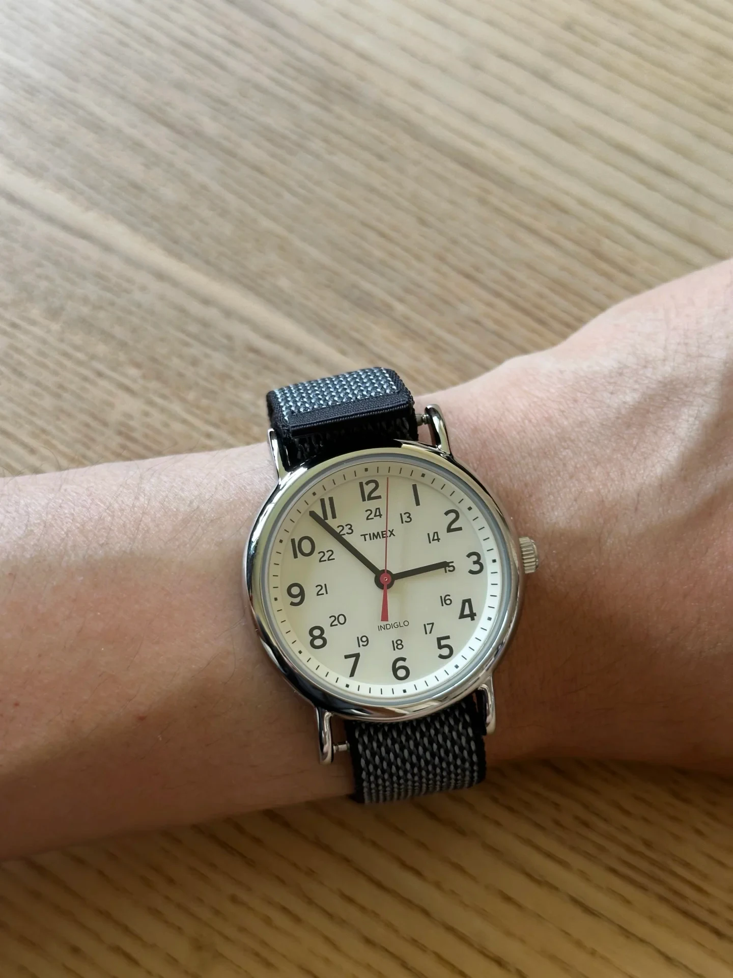 A Timex Indiglo watch with a round silver case, cream dial, Arabic numerals, and a woven patterned strap worn on a wrist against a wooden tabletop.