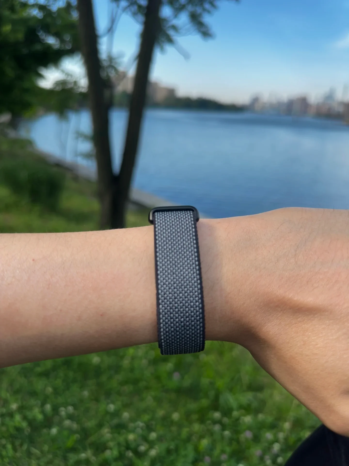 A close-up view of the underside of a wrist wearing a woven dark gray sport loop watch band against an outdoor park and lake background.
