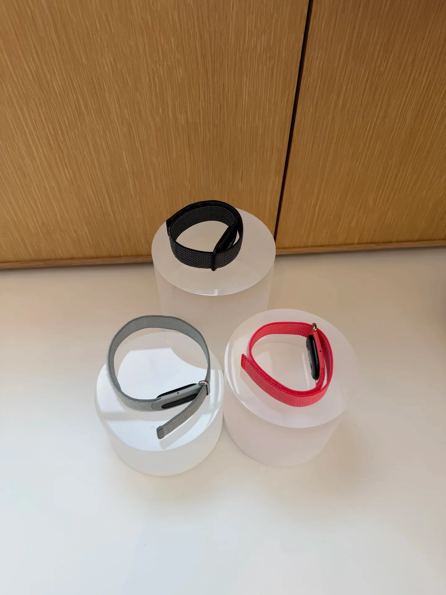 Three fitness bands with fabric straps in black, grey, and red displayed on clear cylindrical acrylic risers in front of a wooden panel.