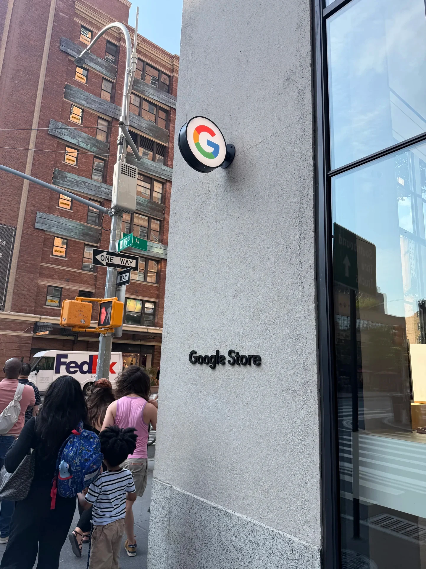 Exterior view of a Google Store with a circular Google logo sign mounted on the building facade, black lettering reading 'Google Store', and pedestrians walking on the sidewalk alongside a street with a FedEx truck.