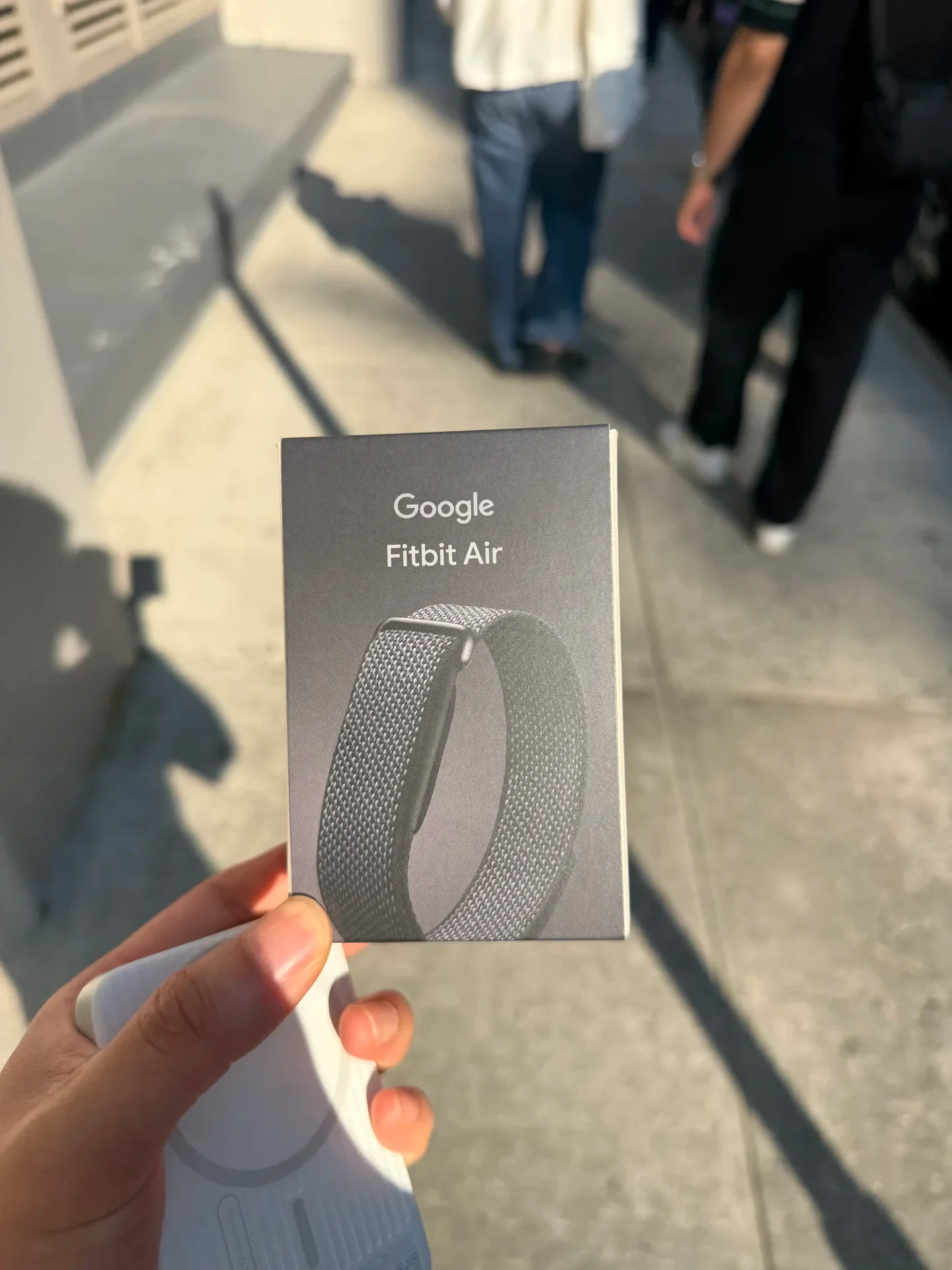 A hand holding a product box for 'Google Fitbit Air' featuring an image of a woven fabric fitness band, outdoors in bright sunlight.