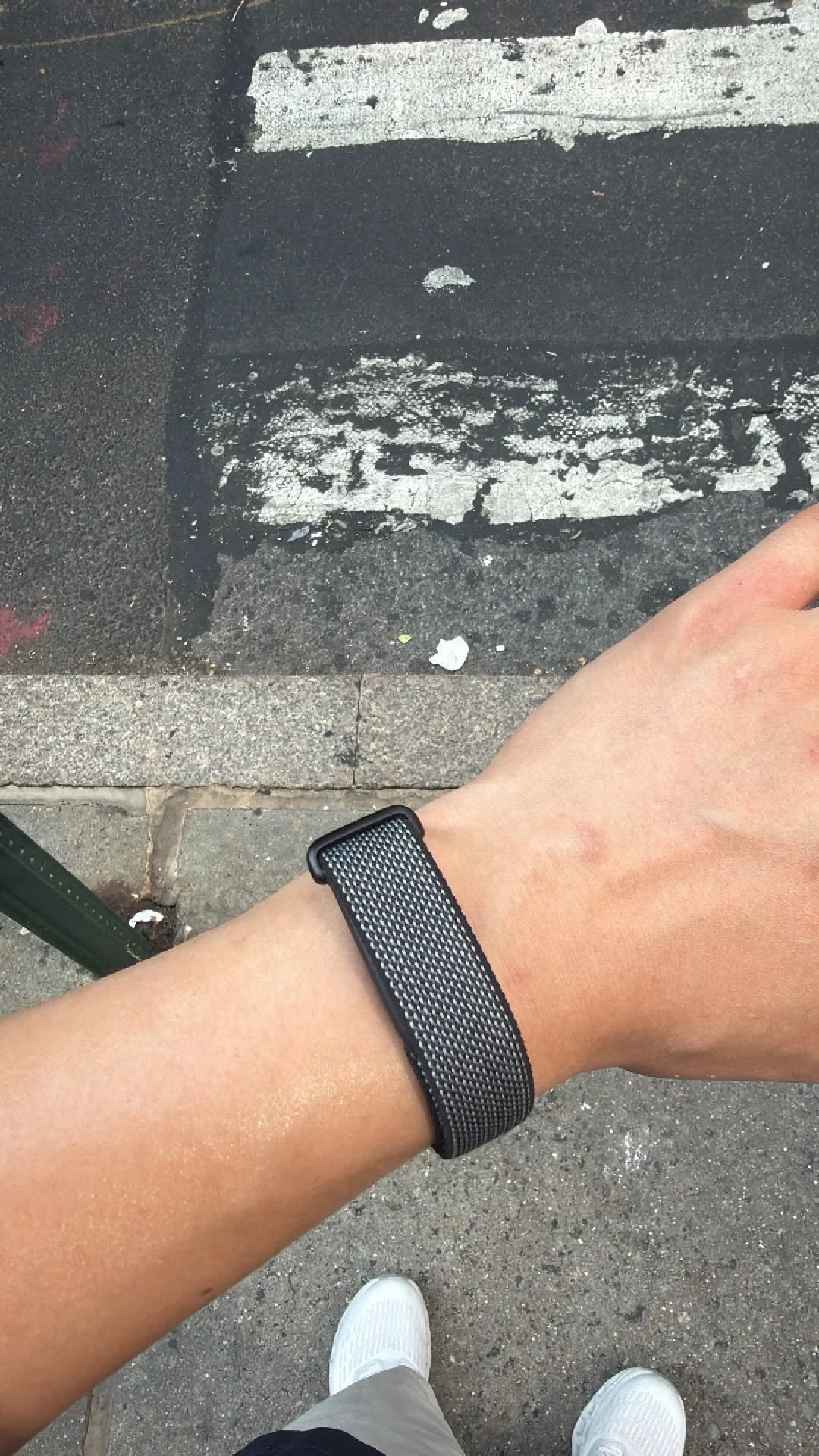First-person view of a wrist wearing a dark gray woven sport loop watch strap while standing outdoors by a crosswalk.